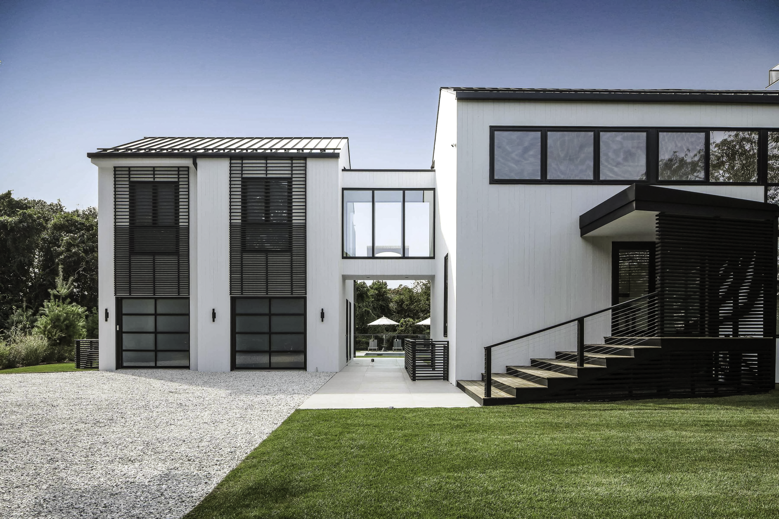 Montauk Interior Design. Modern white house with black accents, windows, and stairs leading to the entrance, green lawn, and gravel driveway under a clear blue sky.