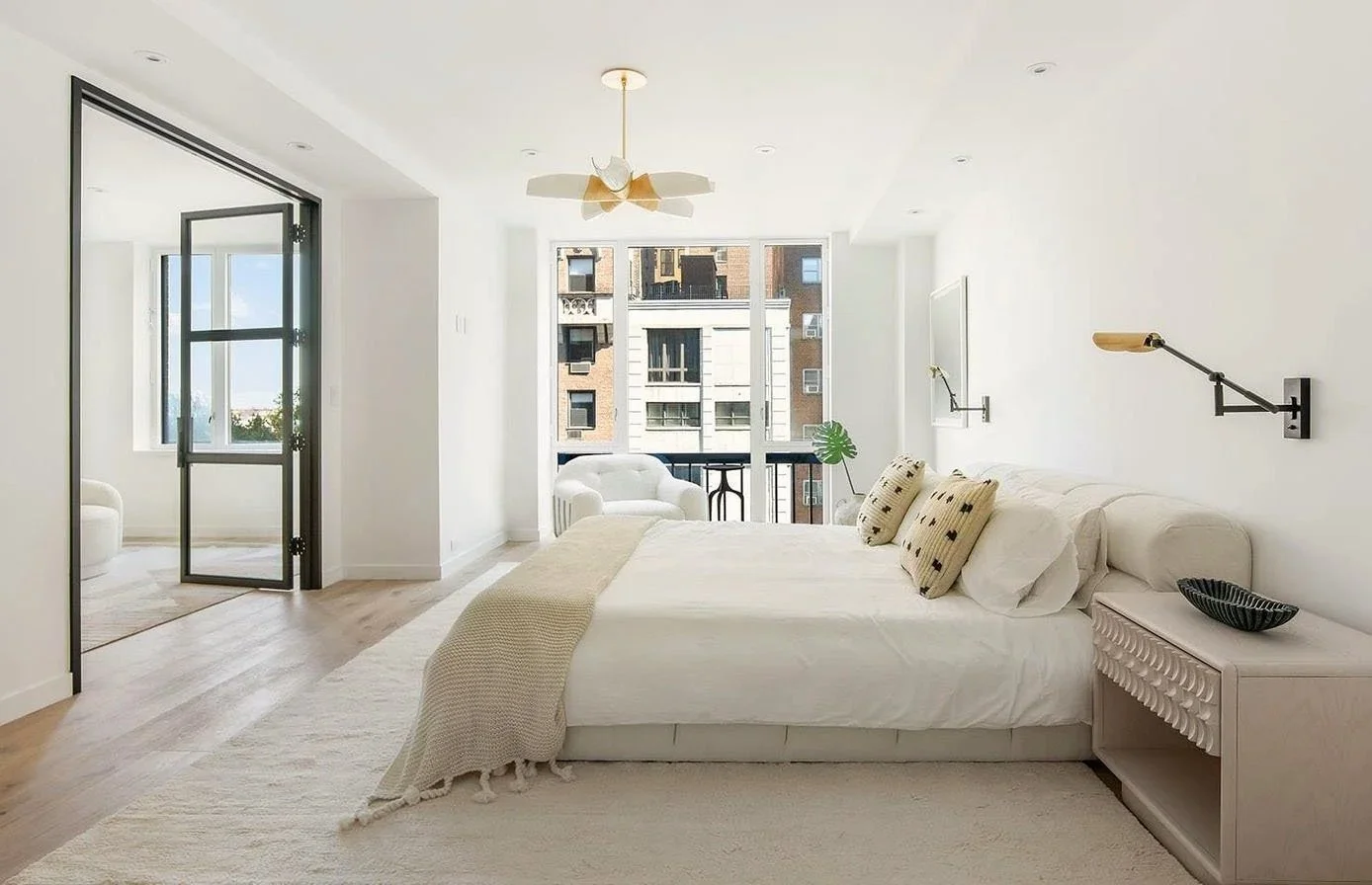 Bright bedroom with large window, white bed, beige blanket, decorative pillows, wall-mounted reading lights, and a small black bowl on the bedside table.