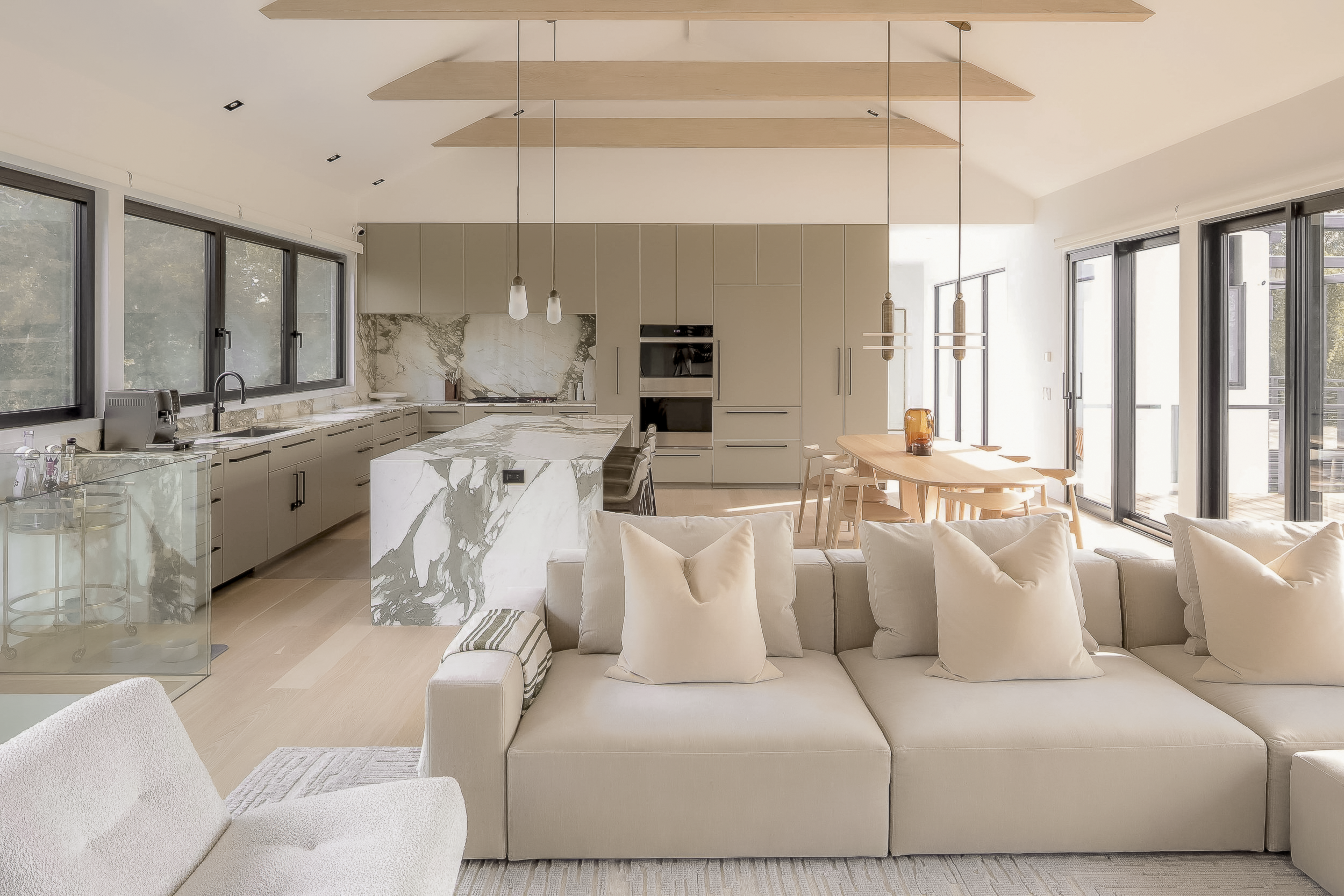 Open concept living space with a kitchen, dining area, and living room. The kitchen features beige cabinets, a marble island, and large windows. The dining area has a wooden table with chairs, and the living room includes a beige sofa with pillows and a light-colored armchair. Sliders and windows allow natural light into the space.