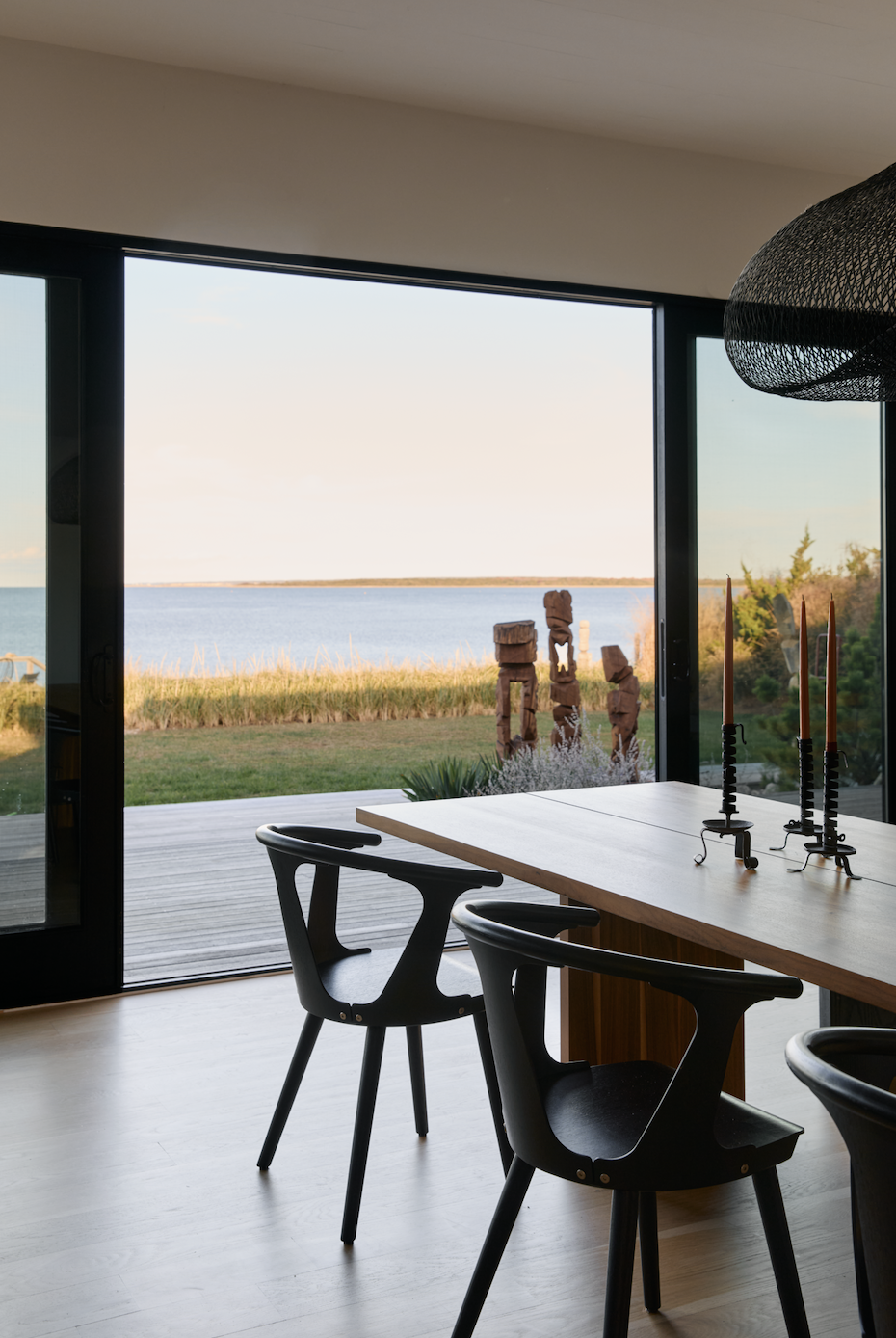 Hamptons Interior Design. Dining room with black chairs and a wooden table near large glass sliding doors, overlooking a body of water with wooden sculptures on the grass outside.