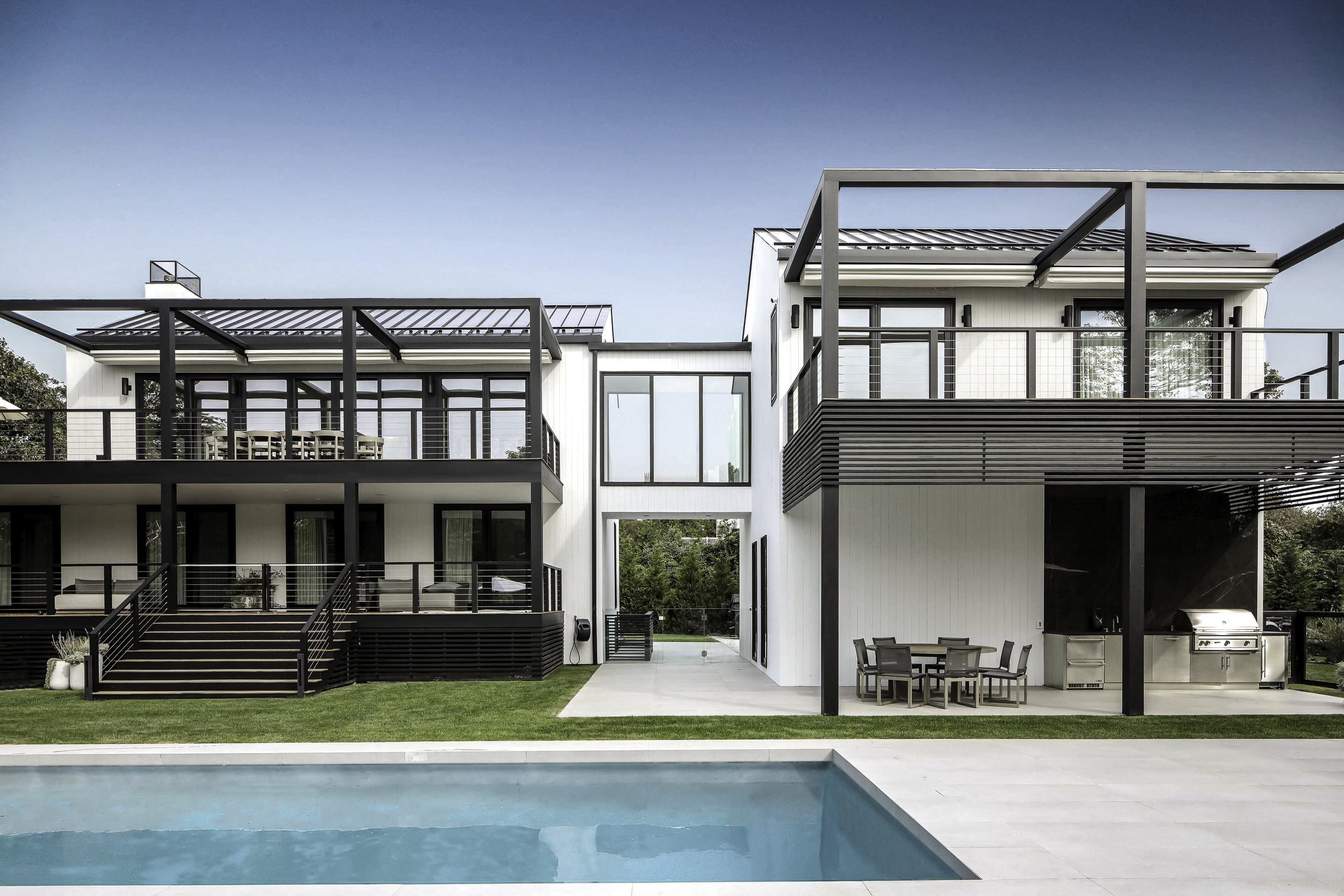 Montauk Interior Design. Modern two-story house with black and white exterior, multiple glass doors and windows, a patio with dining and grilling areas, and a swimming pool in the foreground