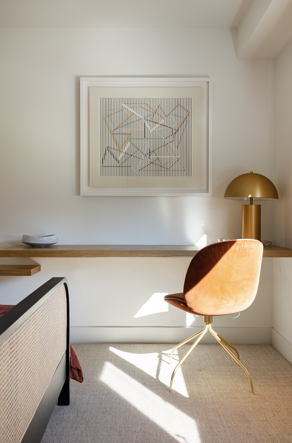 Hamptons Interior Design. A minimalist home office corner with a brown upholstered swivel chair, a wall-mounted wooden desk, a decorative table lamp, and an abstract framed artwork on the wall.