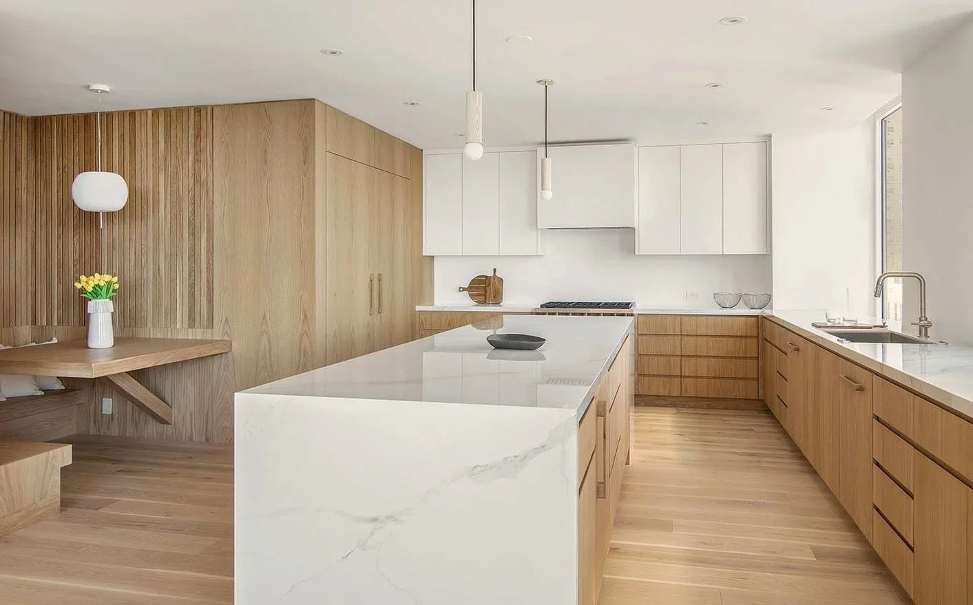 Modern kitchen with light wooden cabinets, a white marble island, white upper cabinets, and a large window with a view outside.