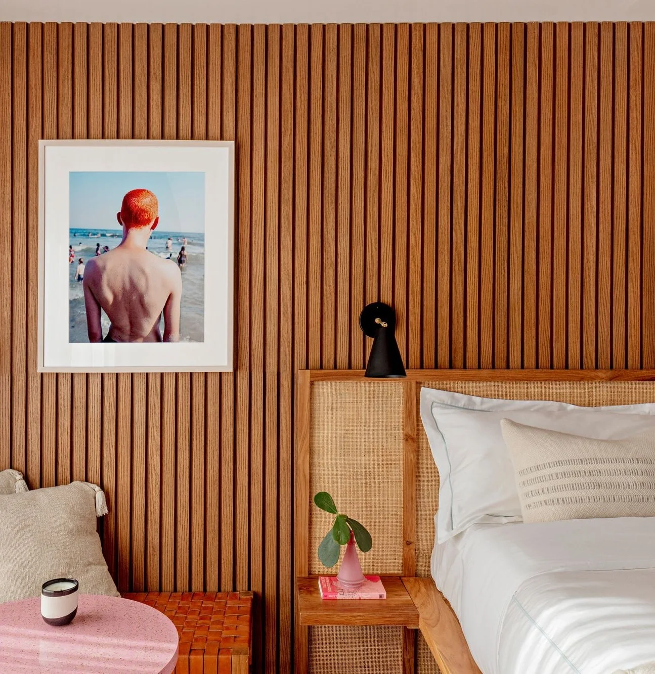 Rockaway Hotel Interior Design. A bedroom with wooden panel walls, a bed with white linens, a wooden side table with a pink vase and green plant, a black wall lamp, a framed beach photograph, and a pink table with a cup and beige cushion.