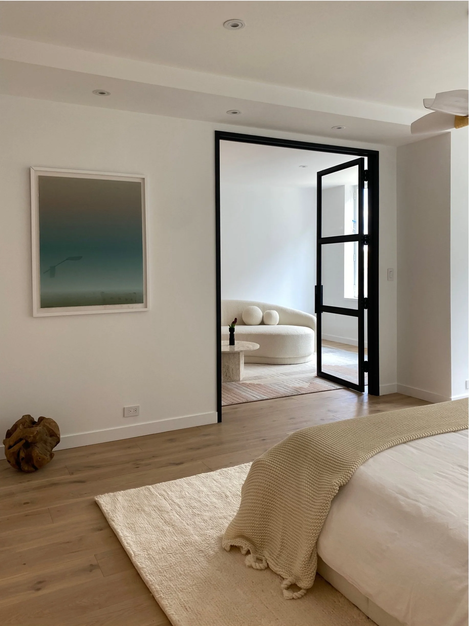 A bedroom with light-colored wooden flooring, a beige bed with a textured blanket, a cream-colored rug, and a wall-mounted mirror. A dark metal-framed glass door opens to a sitting area with a round beige sofa, a small side table with a flower vase, and natural light from a window.