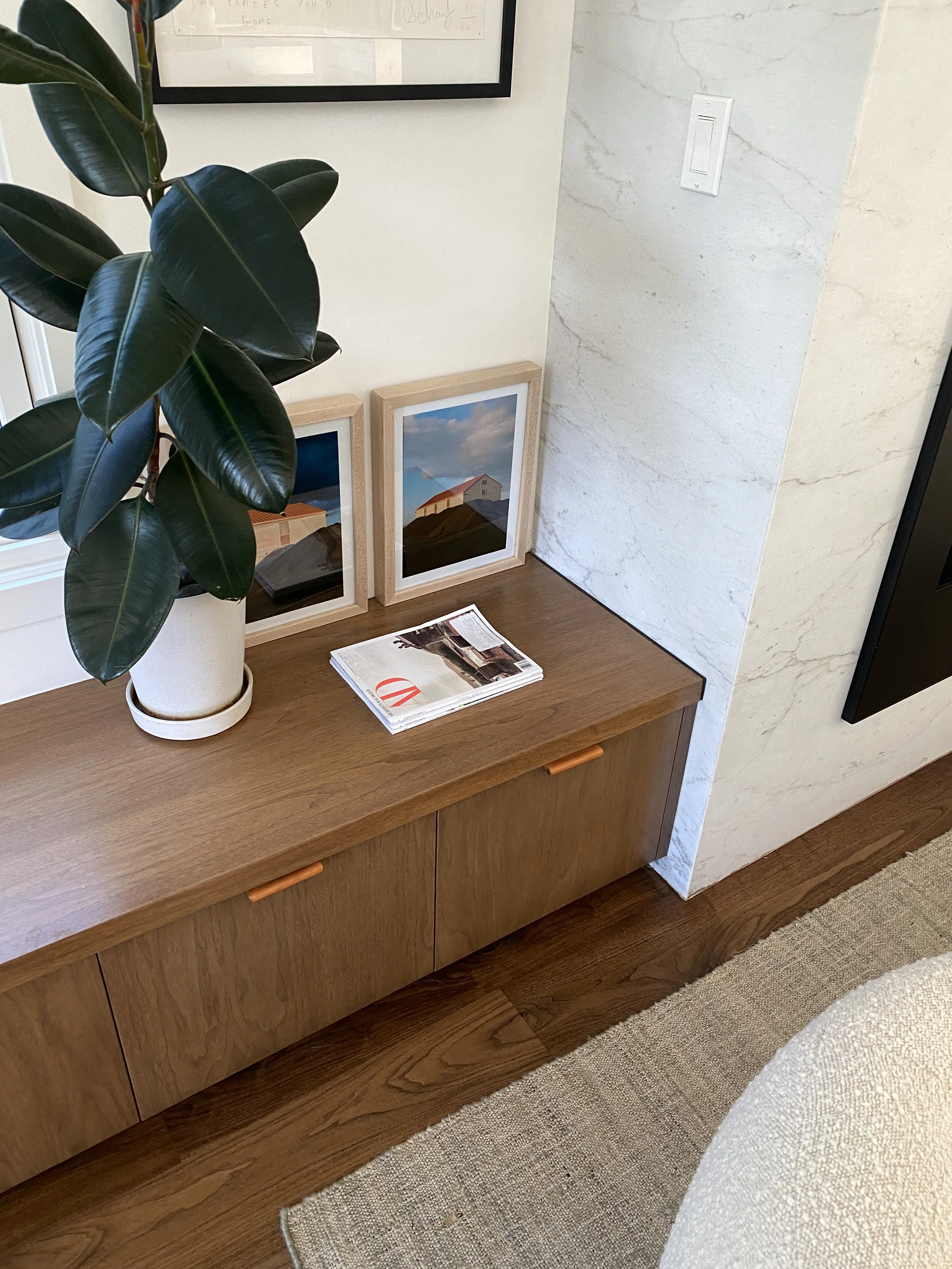 Los Angeles interior Design. A wooden console table holding a potted plant, two framed pictures of houses, and a stack of magazines or brochures, with a white wall, a part of a window with a white frame, and a beige rug nearby.