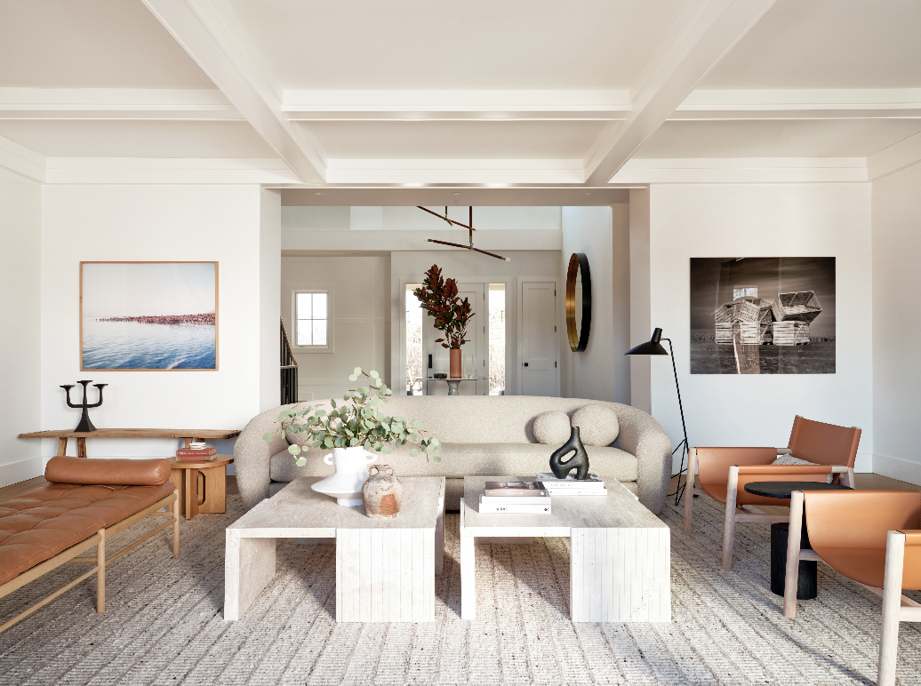 A spacious, modern living room with white walls and ceiling beams, featuring a beige sofa, a wooden bench with leather cushion, two orange chairs, and a coffee table with decorative items. Artwork and a lamp are on the walls.