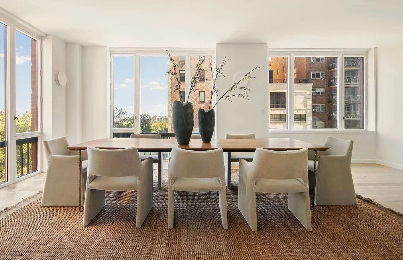 Bright dining room with large windows, white walls, a wooden table, white chairs, and two tall vases with branches as a centerpiece.