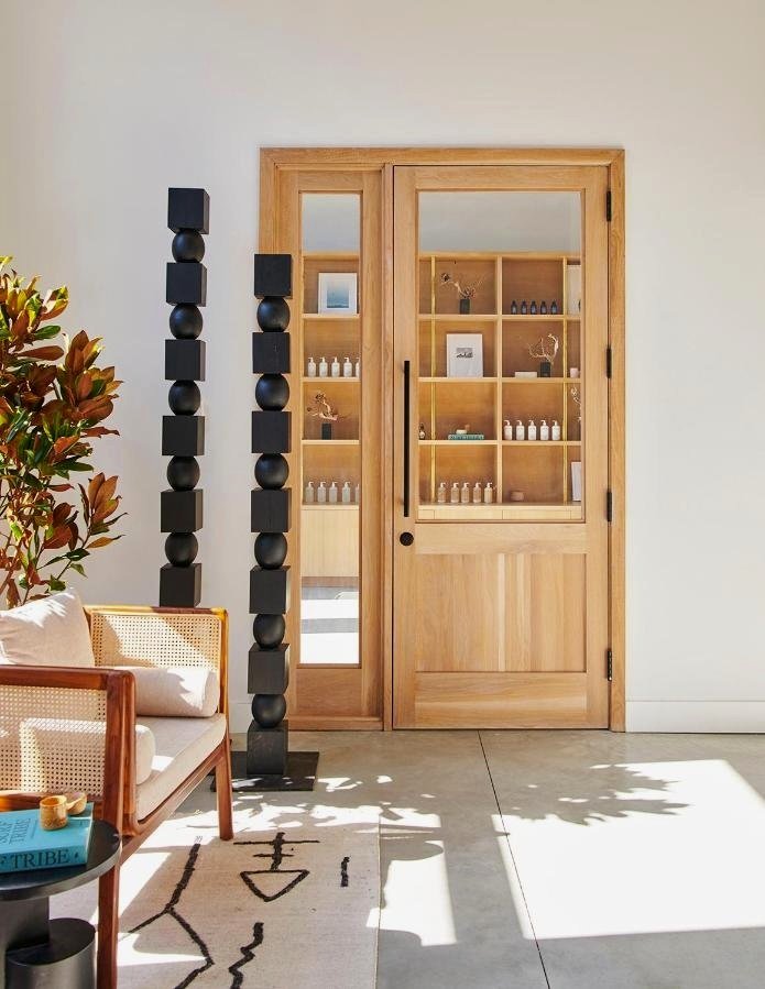 Wooden sliding door with glass panes, interior shelves with decorative items, plants, and framed art, sunlight casting shadows on the floor, part of a sitting area with a rattan chair and a plant.