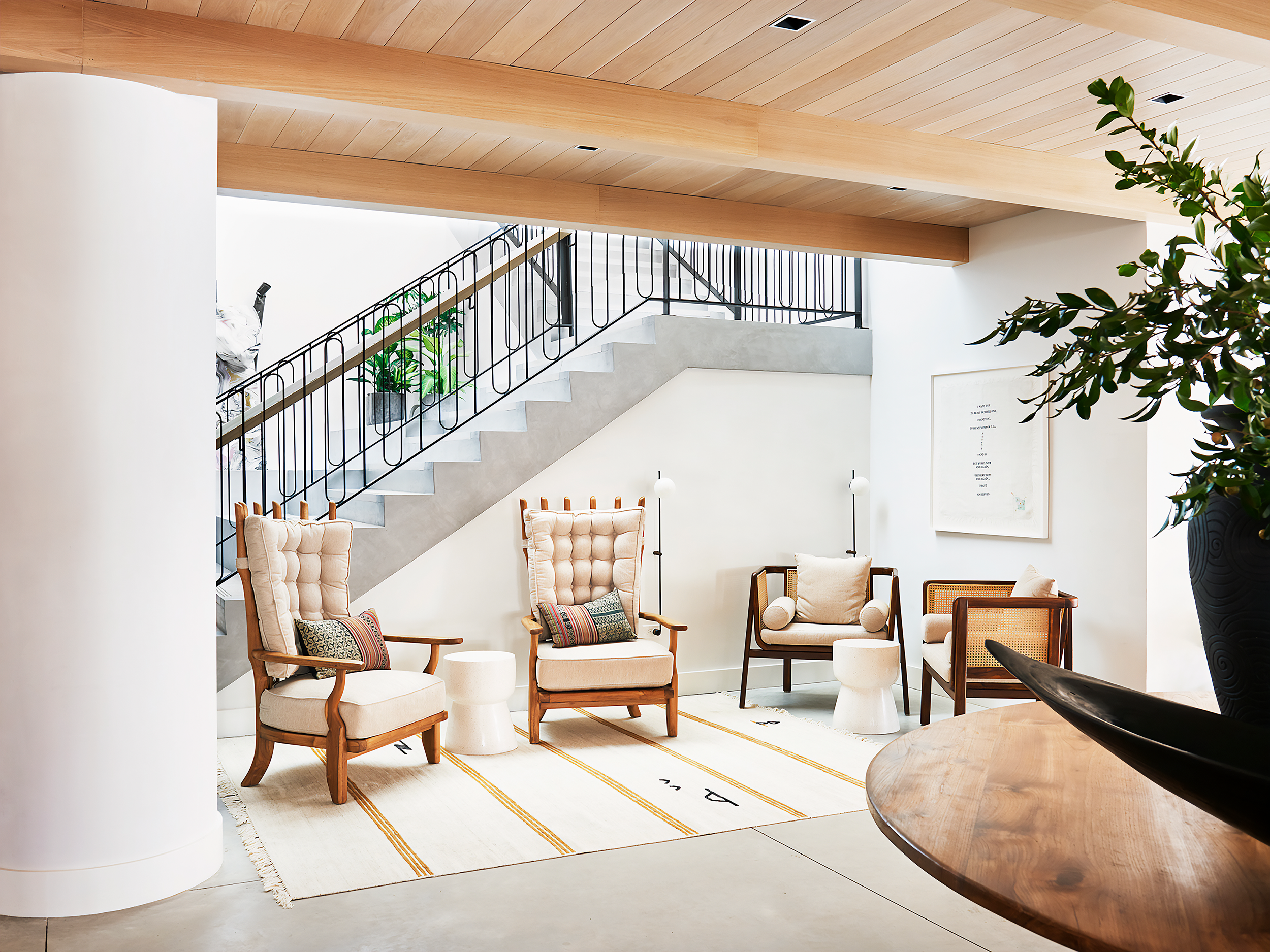 A modern, minimalist living space with four beige armchairs with wooden frames and cushions, small round side tables, a patterned rug with yellow lines, a staircase with black railing and concrete steps, potted plants, and a framed artwork on the wall.