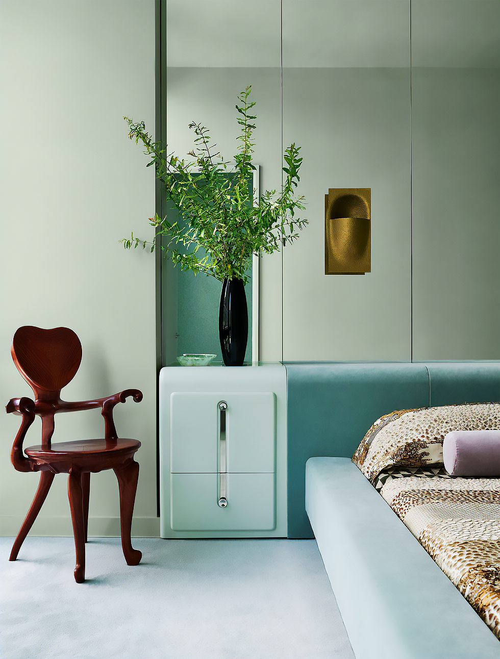 New York Interior Design. Tribeca Design. Interior bedroom with a red stylish chair, a white nightstand with a black vase containing green foliage, a bed with patterned bedding, and a gold wall-mounted light fixture.