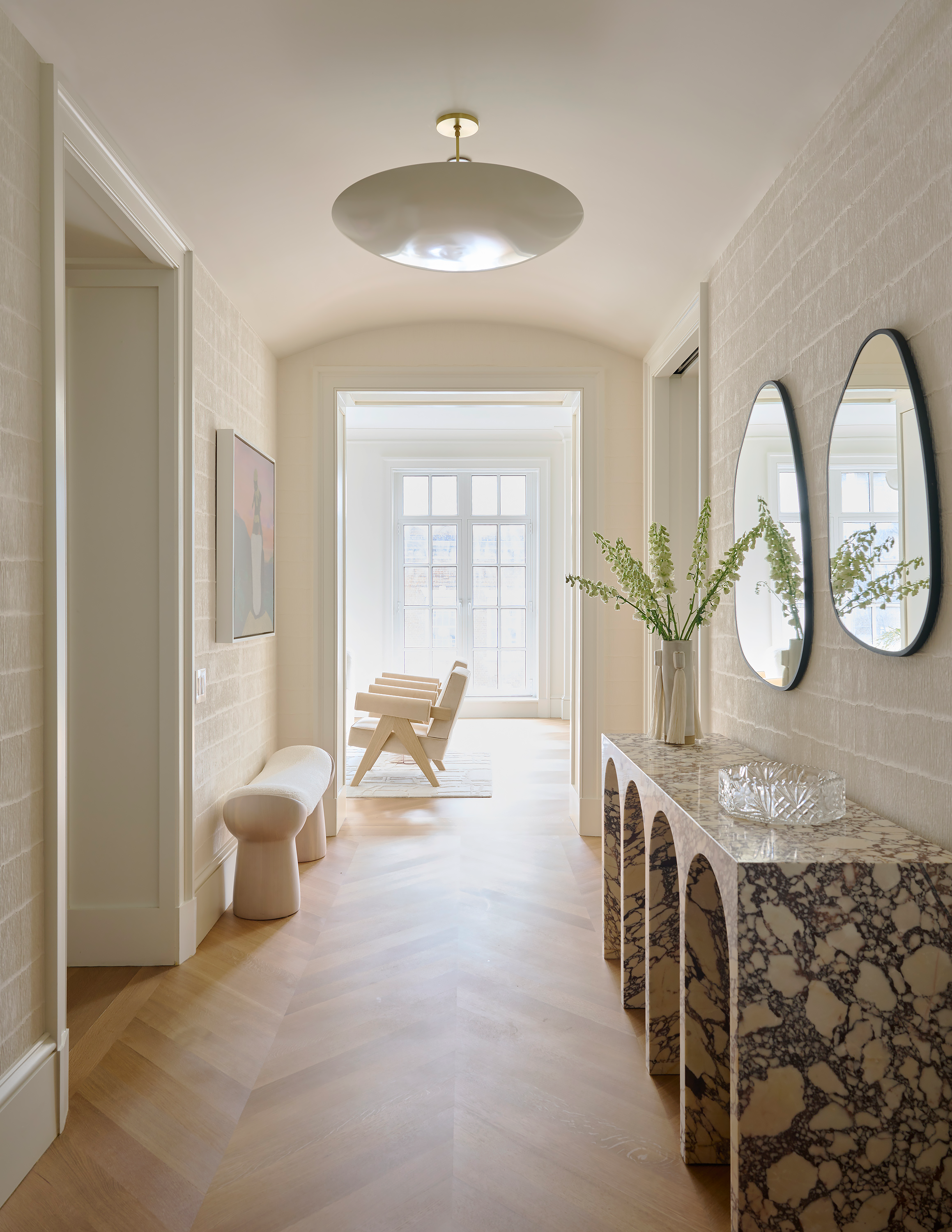 New York Interior Design. Upper East Side interior. Bright hallway with a marble console table, two circular mirrors, a tall white vase with green branches, and a wooden bench. Large window at the end of the hall allows natural light.
