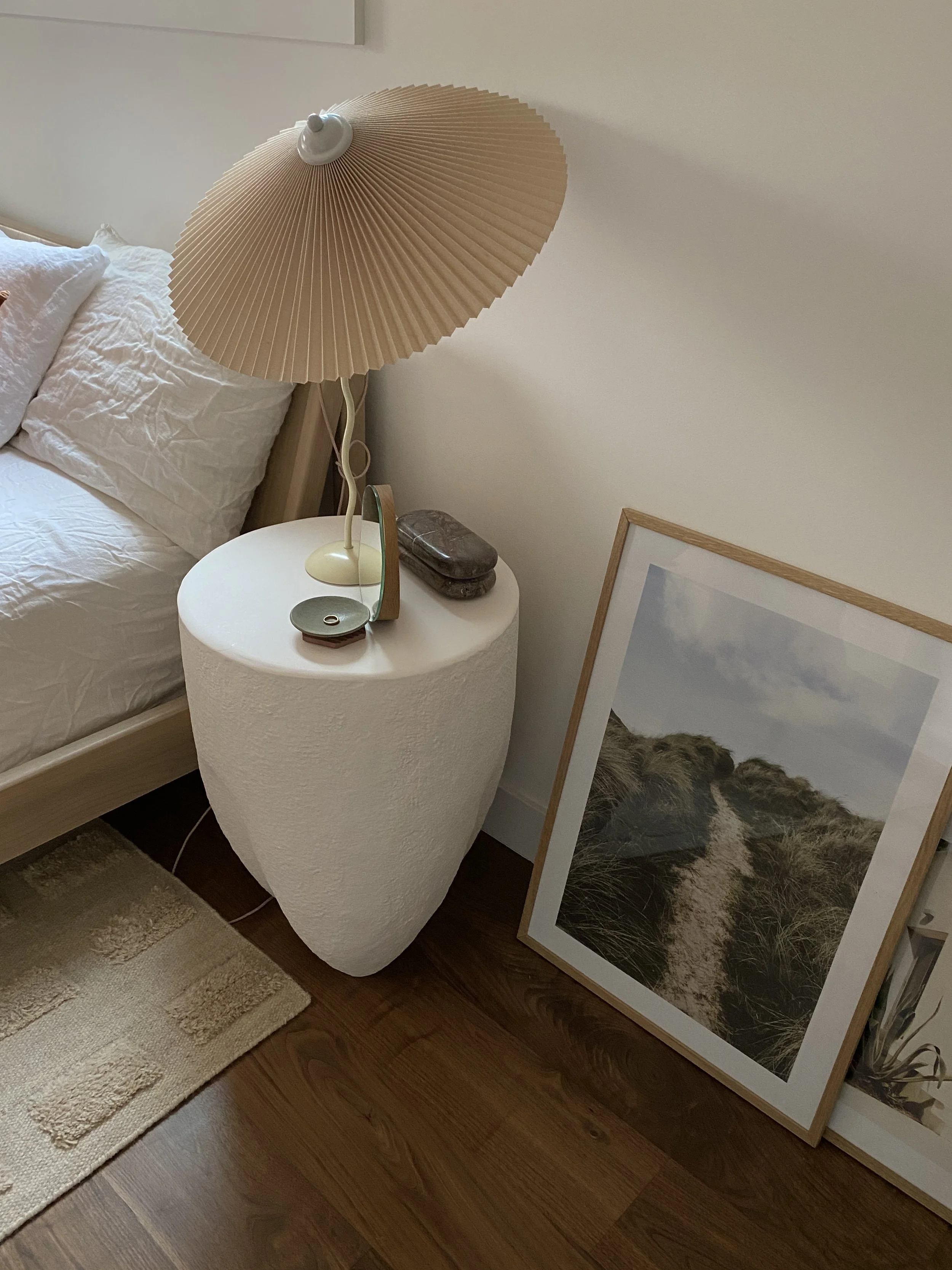 A bedroom corner featuring a bed with white sheets and pillows, a beige bedside table with a table lamp, a decorative object, and a small stone object, and a large framed photo of a sandy pathway through grassy dunes leaning against the wall.