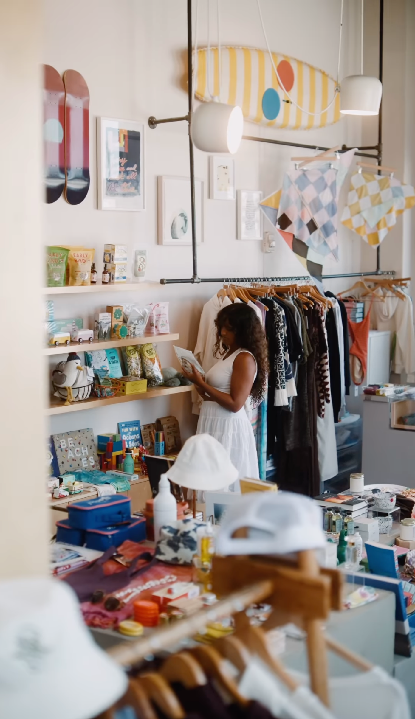 A woman shopping in a boutique store filled with clothing, toys, and accessories. She is wearing a white dress and has long curly hair, browsing items on a shelf.