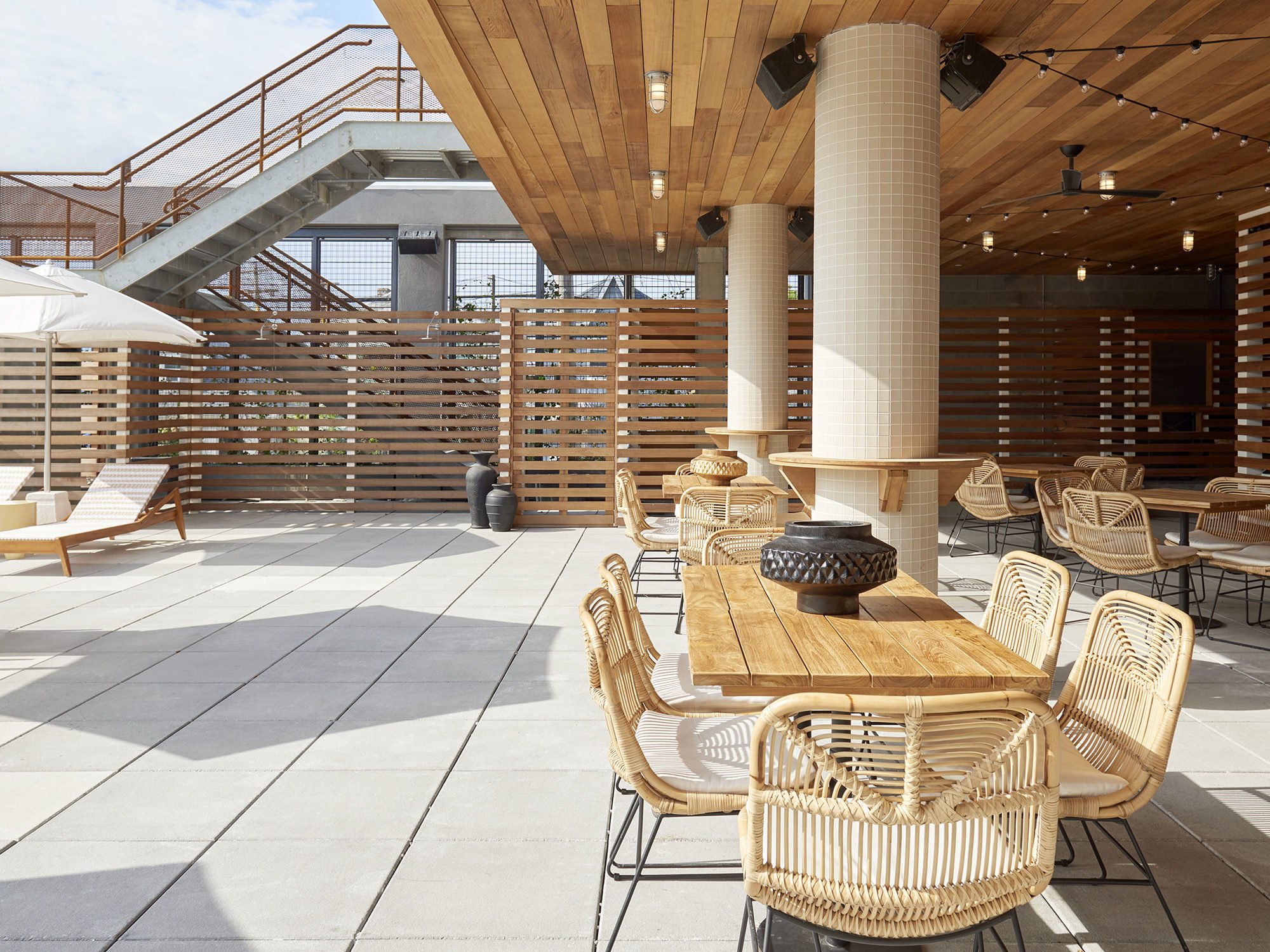 Outdoor rooftop seating area with wooden tables, wicker chairs, a lounge chair, and decorative vases, under string lights and a wooden ceiling.
