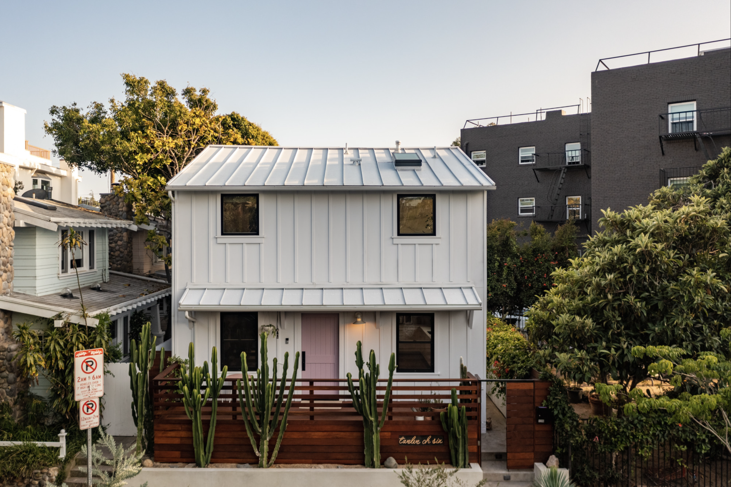 Los Angeles interior Design. A modern, white two-story house with a pink front door, metal roof, and surrounded by green plants and cacti. There is a wooden fence in front and a sign indicating parking restrictions.