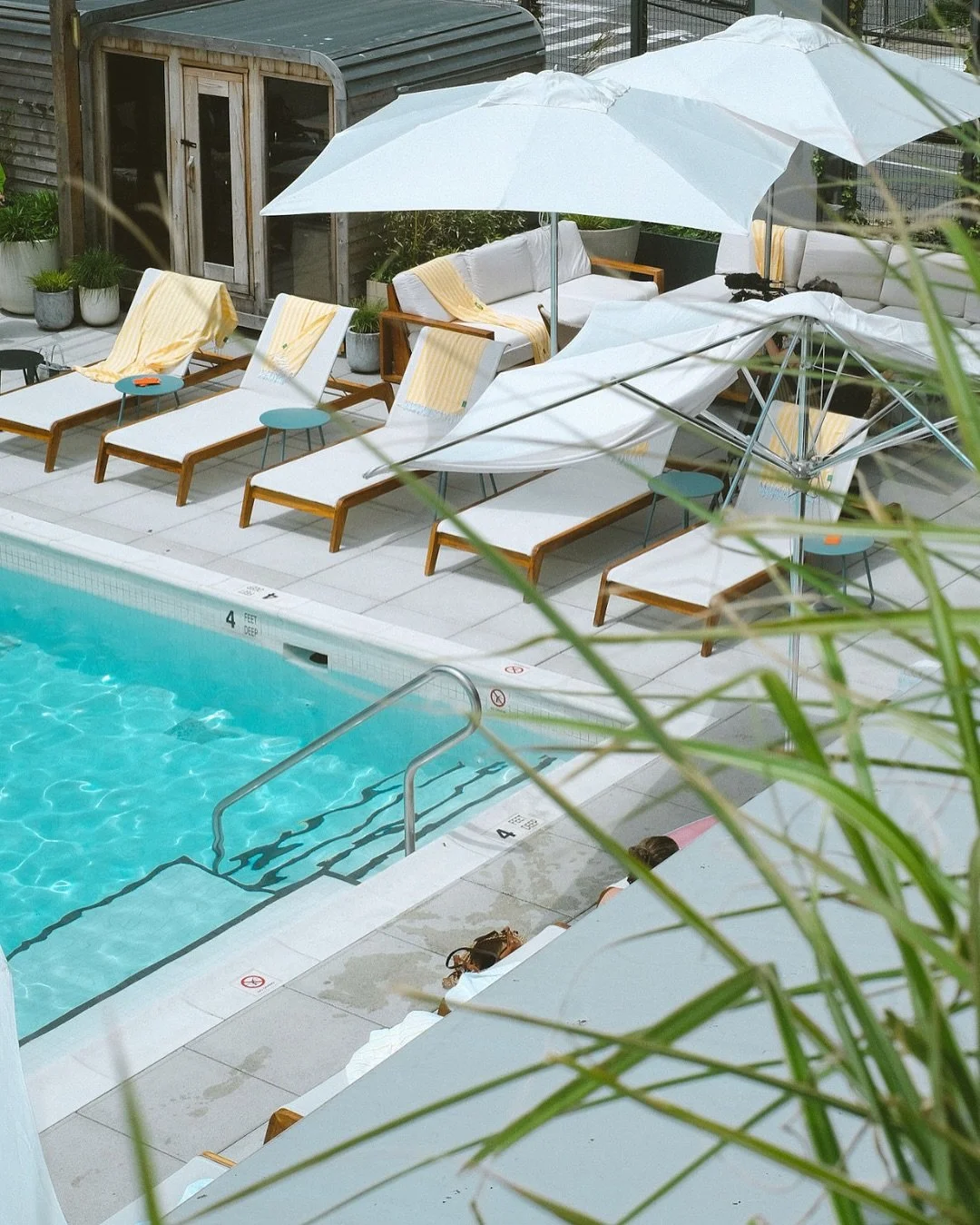 Empty poolside area with white lounge chairs, yellow towels, blue side tables, large white umbrellas, potted plants, and a dog house in the background. One person is partially visible near the pool.