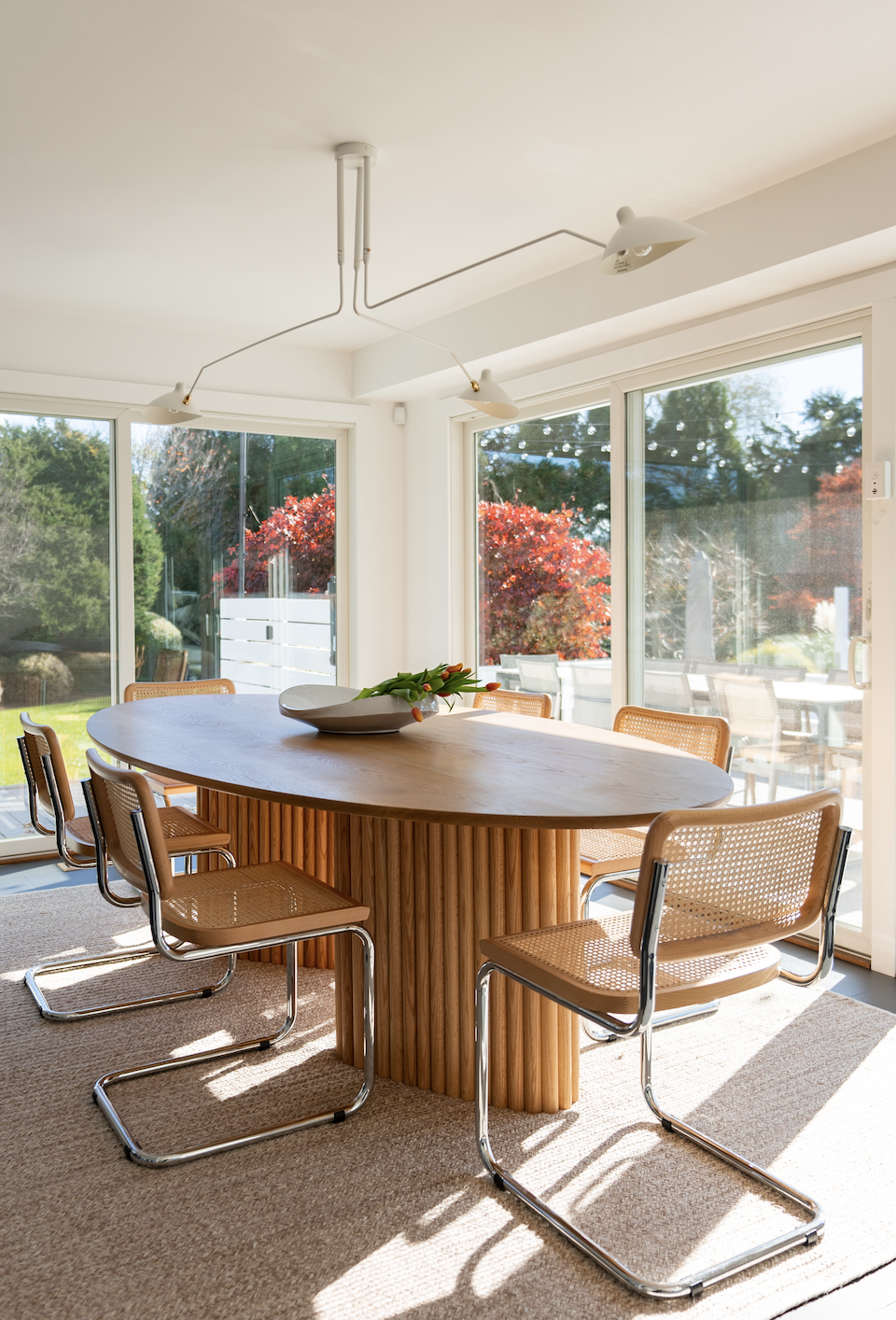 Hamptons Interior Design. Bright dining room with a wooden oval table, six cane and metal chairs, and large windows showing a garden with trees and red foliage.