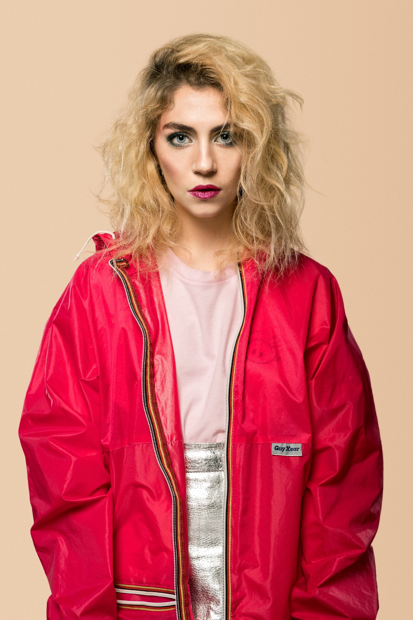 Femme aux cheveux blonds portant une veste rouge et un t-shirt rose sur un fond beige.