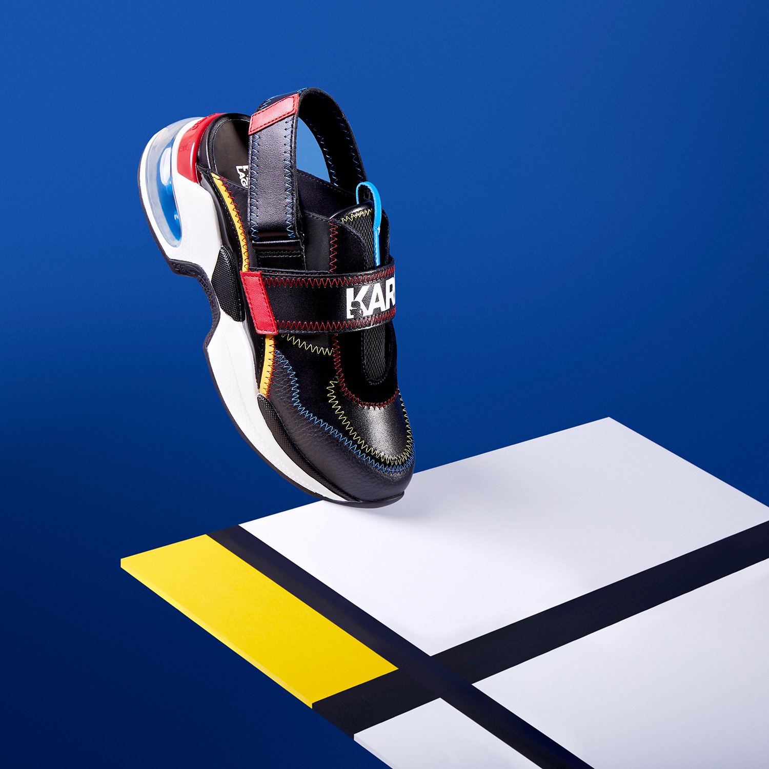 Chaussure de sport noire avec détails colorés, semelle épaisse, sur fond bleu avec motifs géométriques jaune, noir et blanc.
