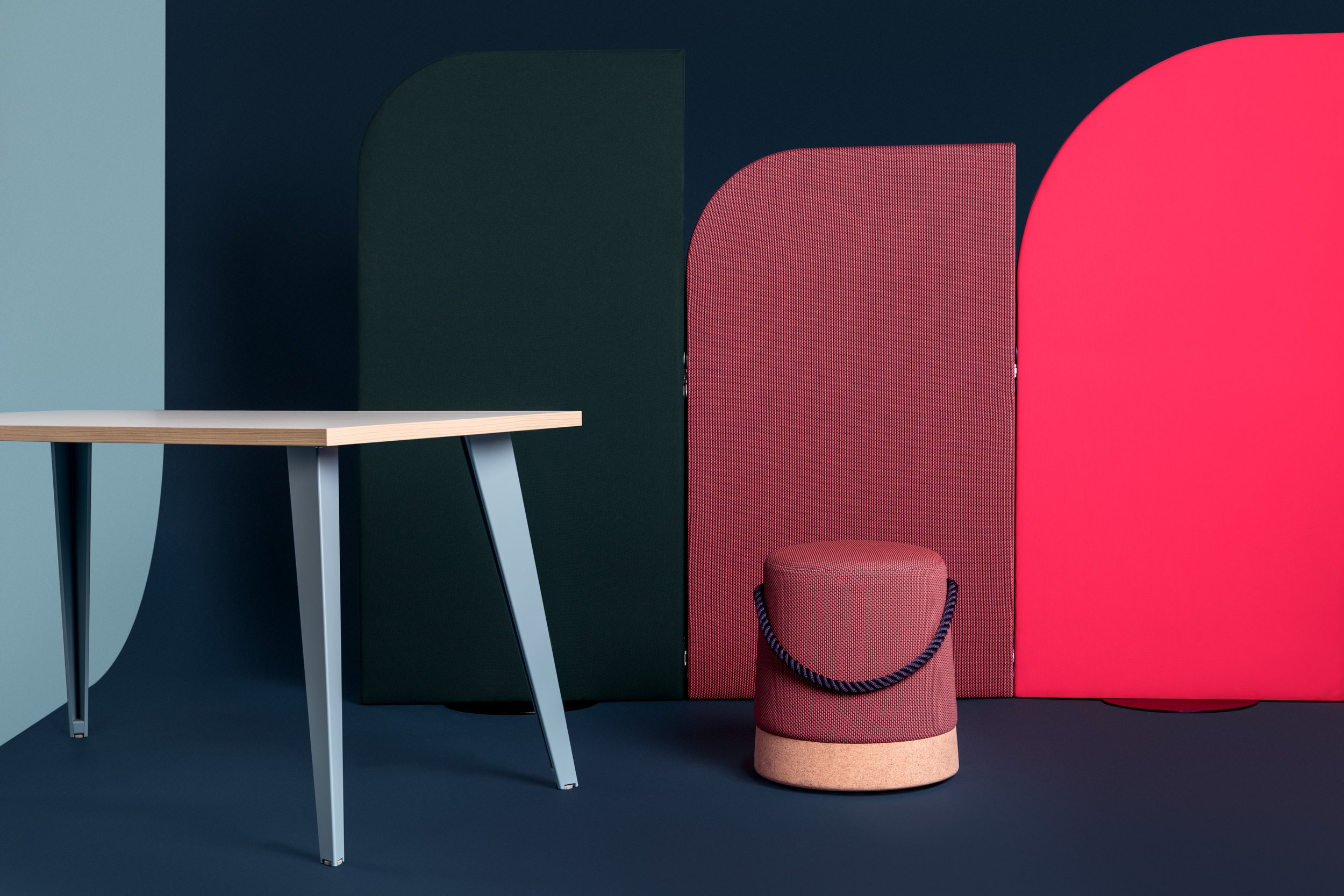 Intérieur moderne avec une table en bois aux pieds métalliques, un petit tabouret rouge avec une base en liège et un cordon, et des parois décoratives colorées en arrière-plan.