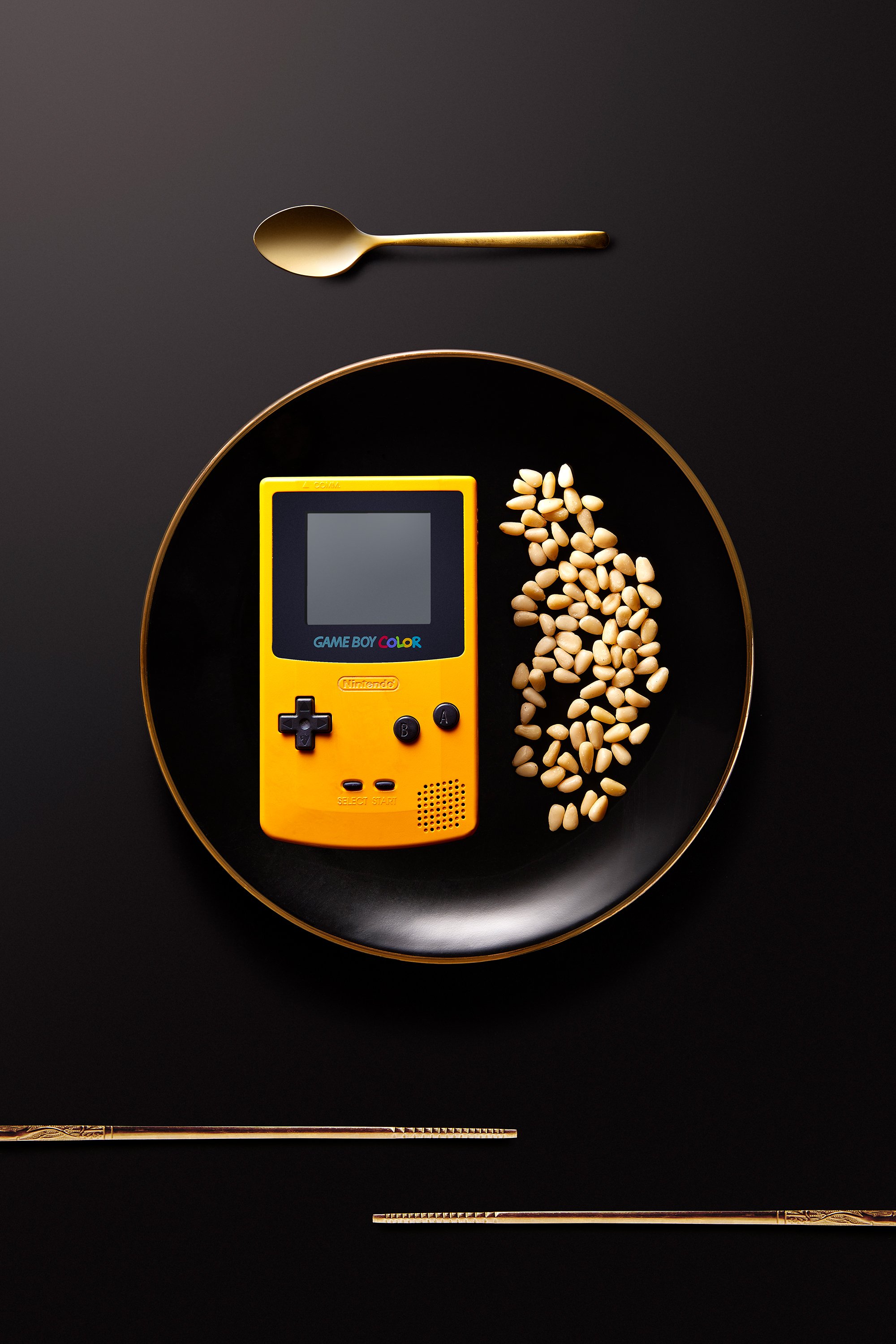 Game Boy Color jaune sur une assiette noire avec des pignons, cuillère et baguettes en or en arrière-plan sur fond noir.