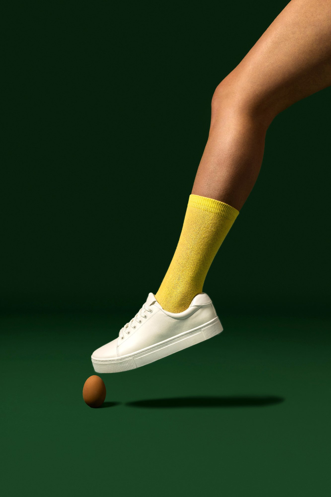 Jambe en chaussette jaune et basket blanche flottant au-dessus d'un œuf brun sur fond vert.