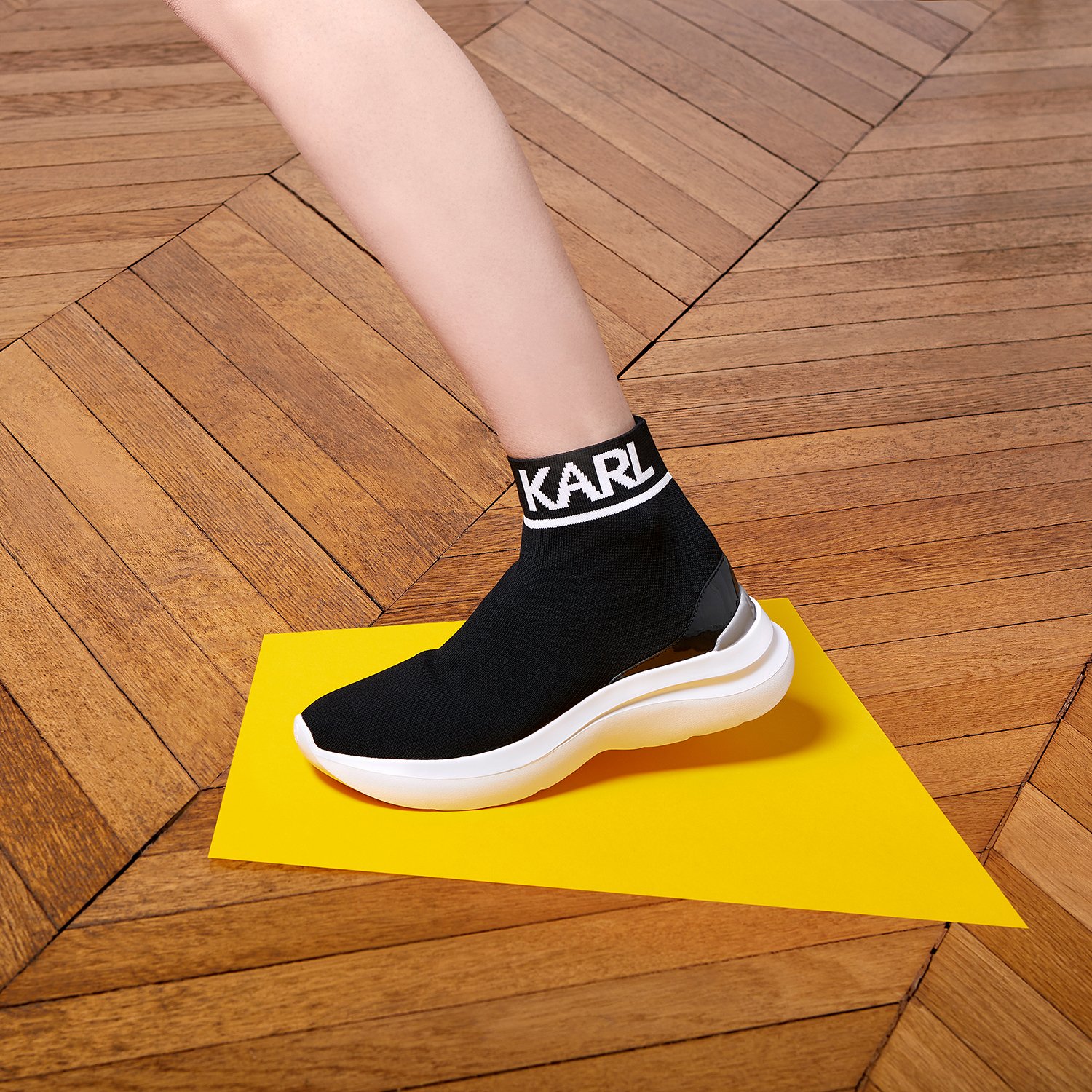 Jambe avec basket noire "KARL" sur sol en bois et triangle jaune