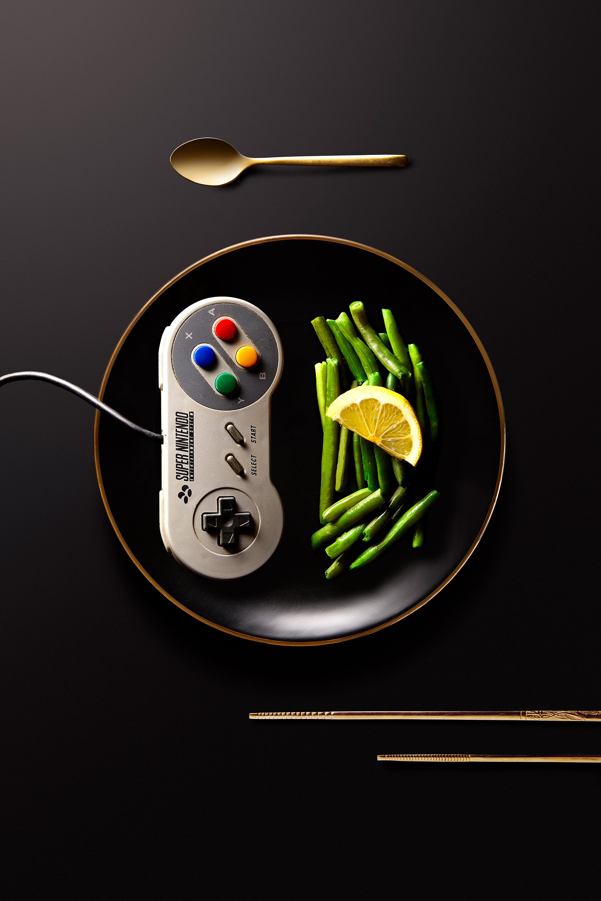 Une manette de jeu Super Nintendo, des haricots verts et une tranche de citron sur une assiette noire, avec une cuillère et des baguettes dorées sur fond noir.