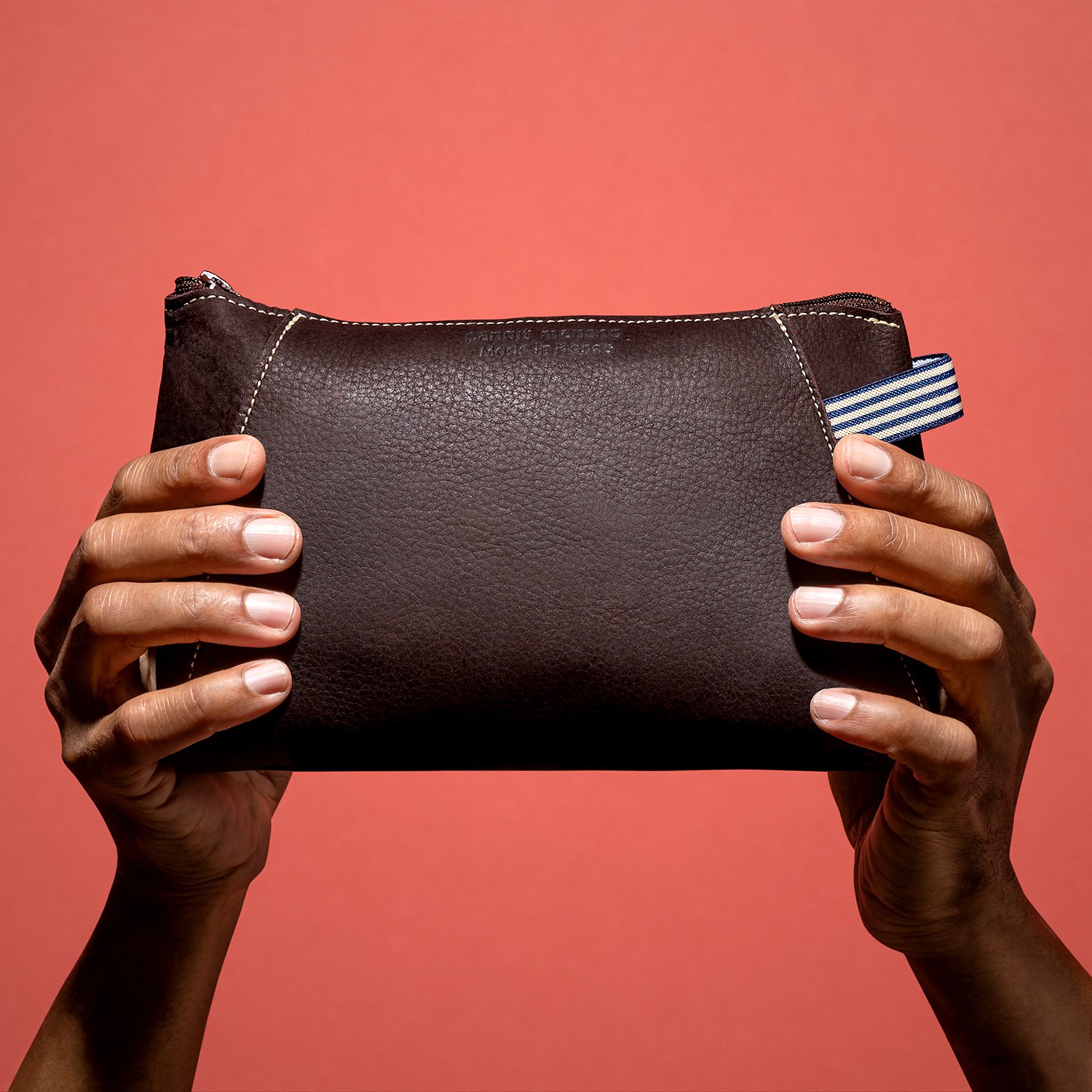 Deux mains tenant une pochette en cuir marron sur fond rouge.