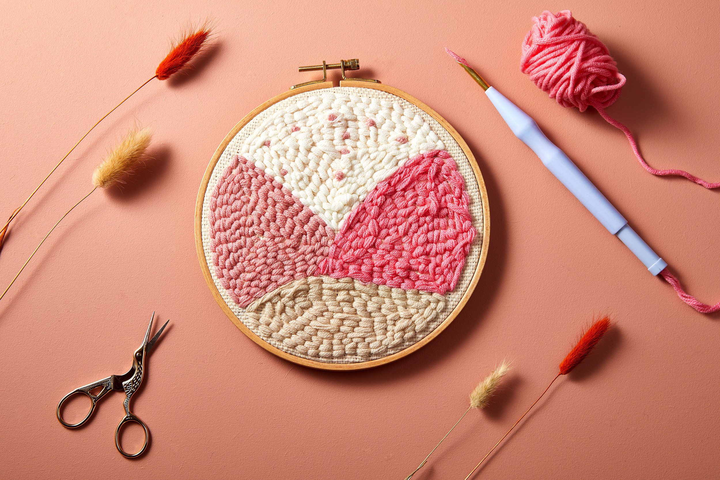 Broderie circulaire dans un cercle à broder en bois avec fils colorés sur fond rose, entourée d'herbes séchées, de ciseaux décoratifs et d'un outil de punch needle avec fil rose.