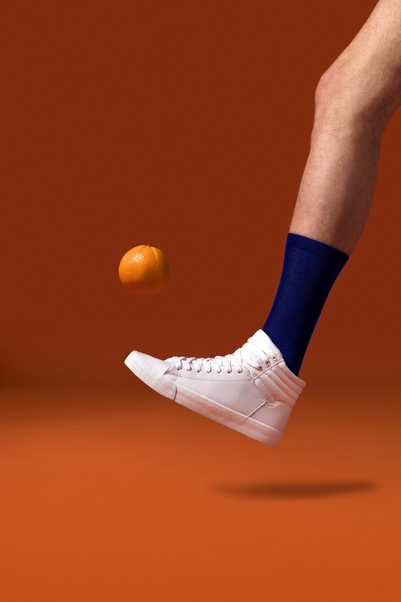 Jambe avec chaussette bleue et basket blanche frappant une orange sur un fond orange.