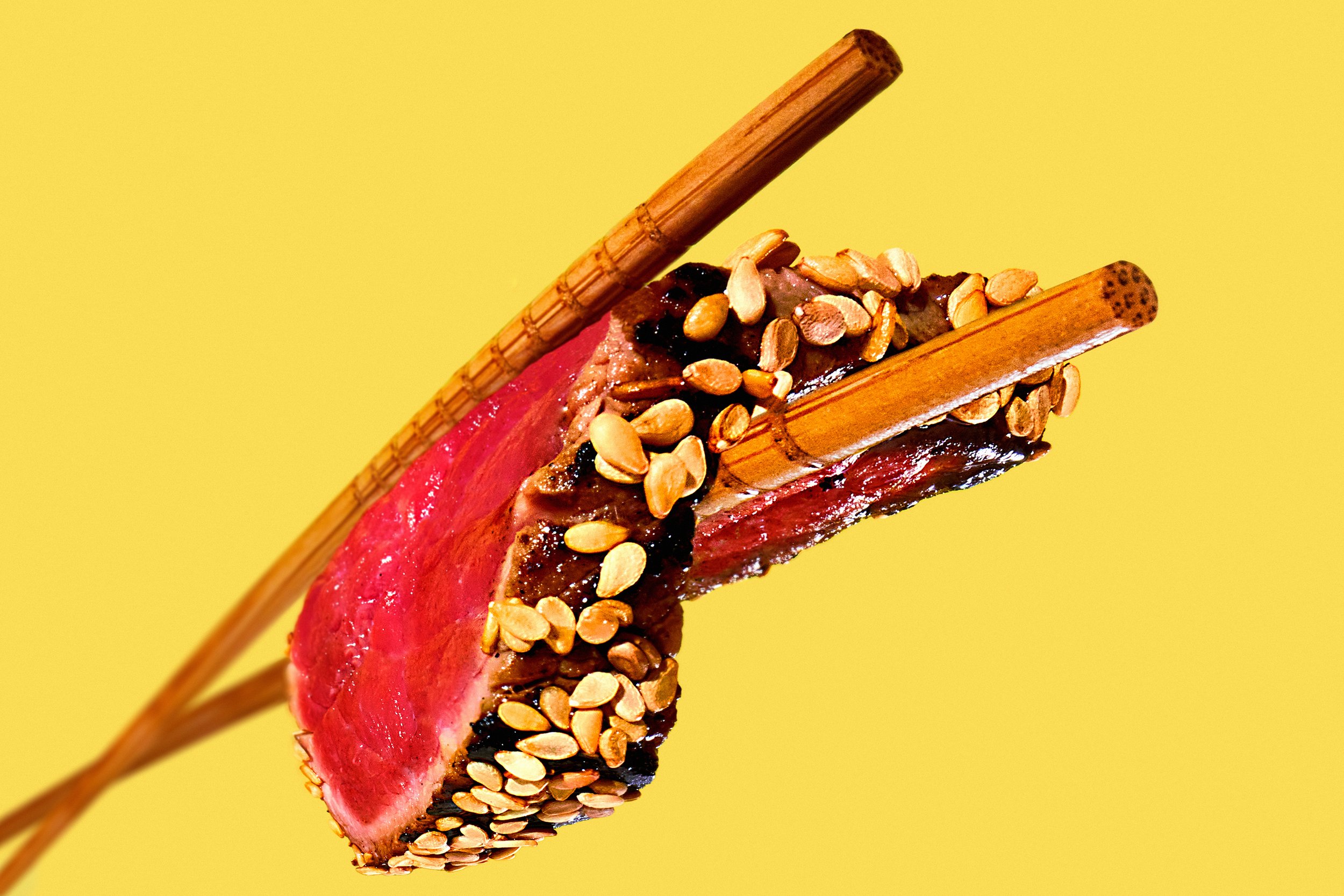 Un morceau de viande rouge avec des graines de sésame, tenu par des baguettes en bois sur un fond jaune.