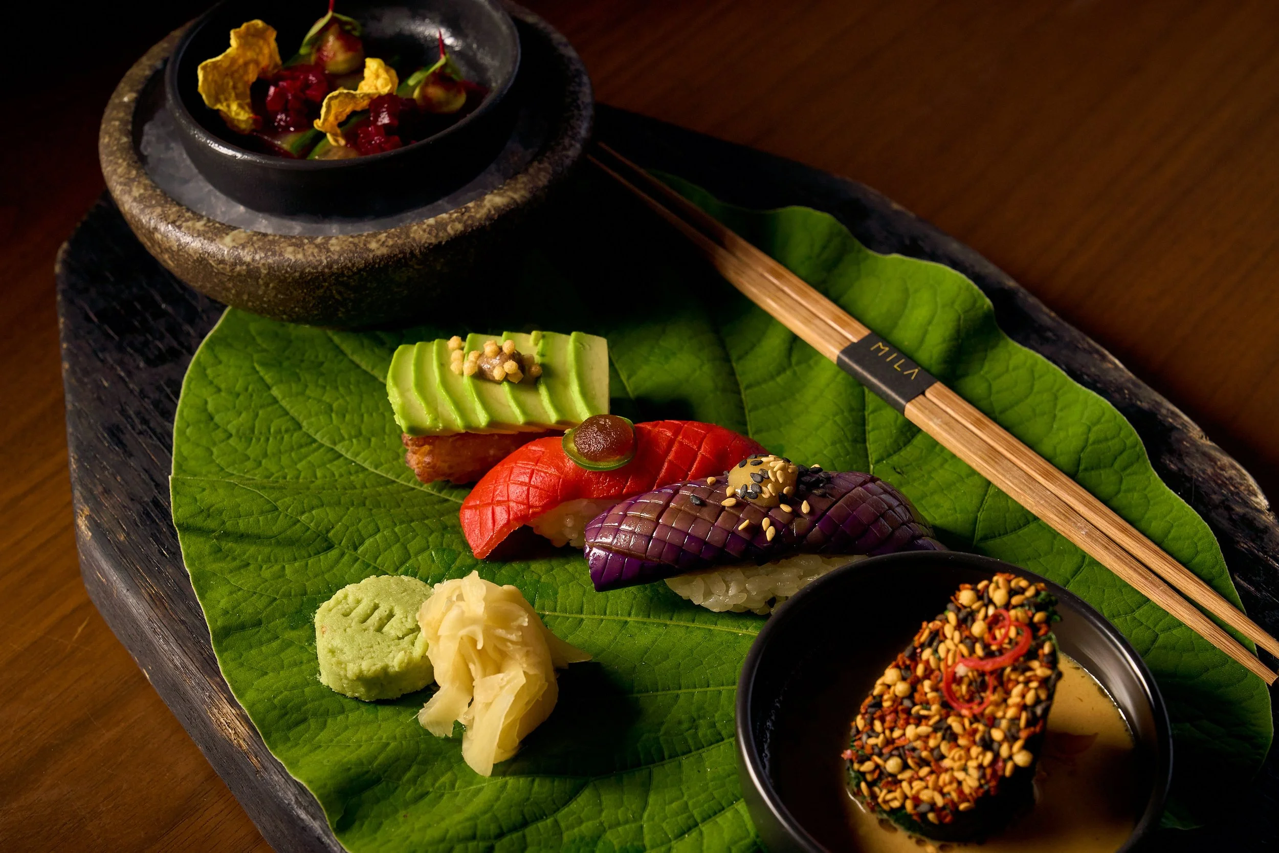 A couple of intricate three sushi's on a leaf, plated with ginger, wasabi and two small dishes.