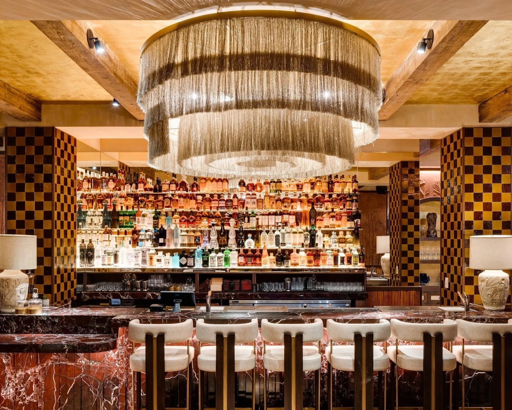 Fully stacked bar with a luxurious decor including a big chandelier, chairs, lamps and marble table tops.