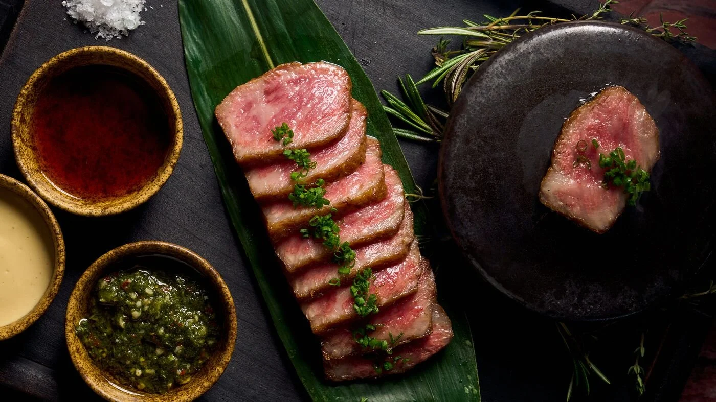 Sliced rare steak on a green leaf with three dipping sauces in small bowls, one green chunky, one dark, and one light. A piece of steak on a dark plate garnished with green onions in the top right corner.