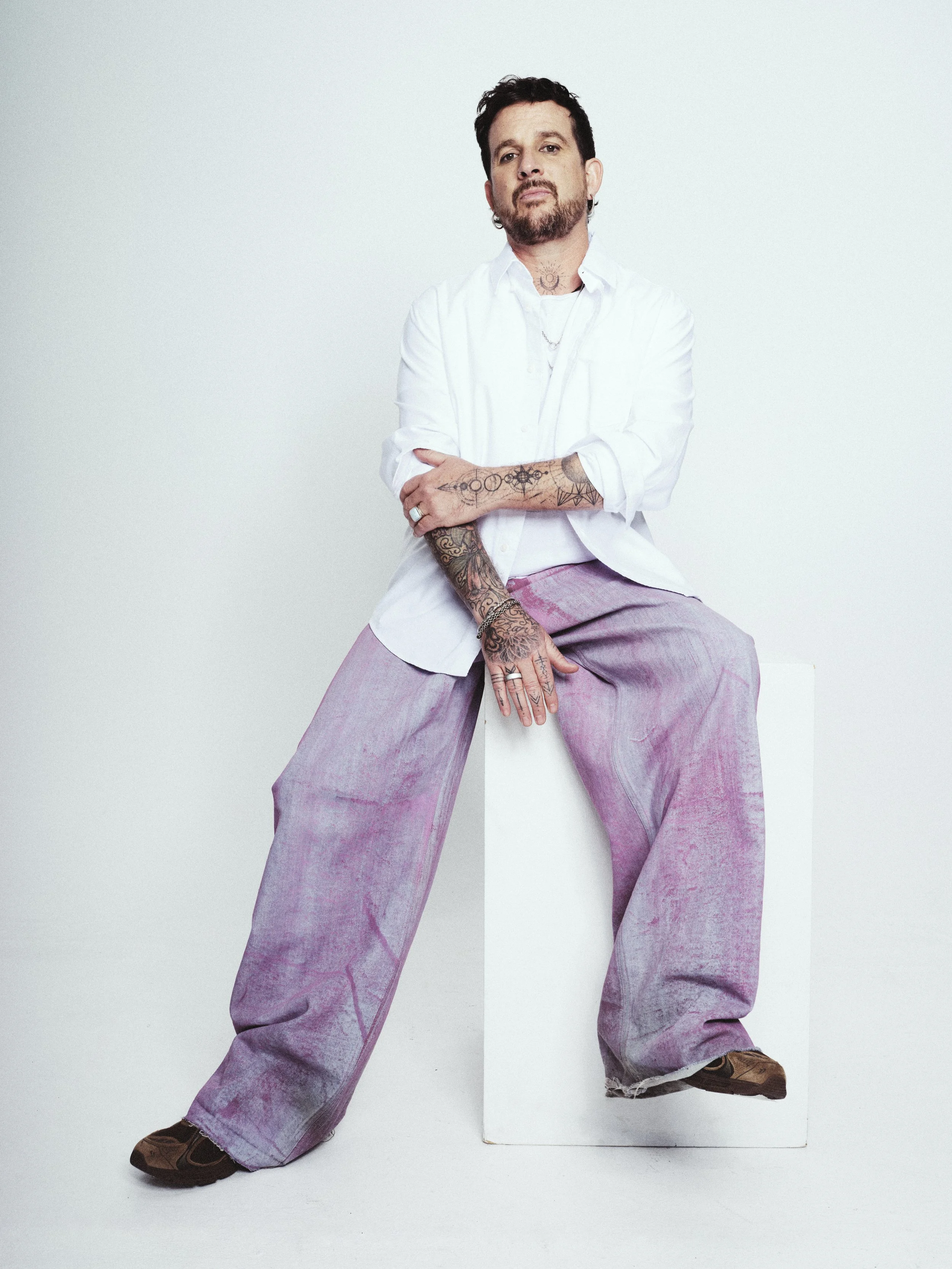 A person with tattoos on arms and chest, wearing a white shirt and pink wide-leg pants, sitting on a stool against a plain background.