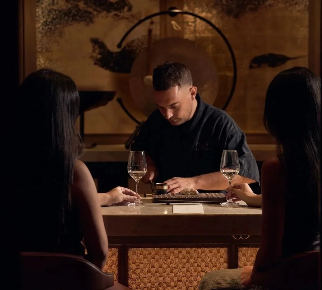 A chef presents a dish at a restaurant table with two women sitting across from him, each with a glass of white wine, in a dimly lit setting with artistic wall decor.