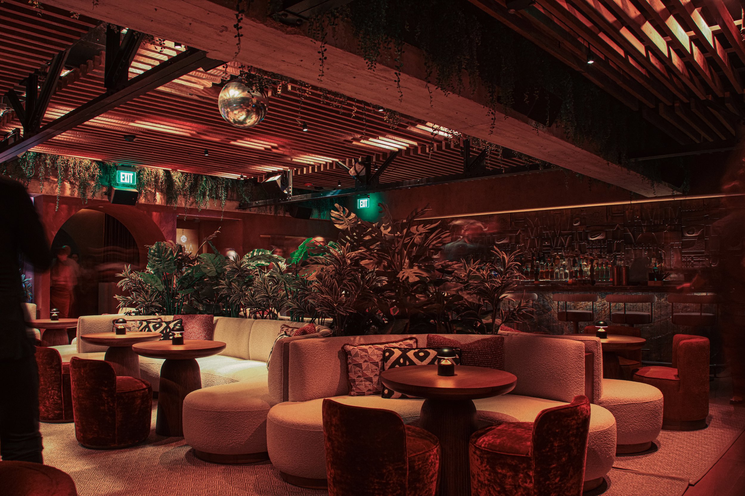 Indoor lounge area with couches, round tables, plants, a wooden ceiling with lighting, and an exit sign.