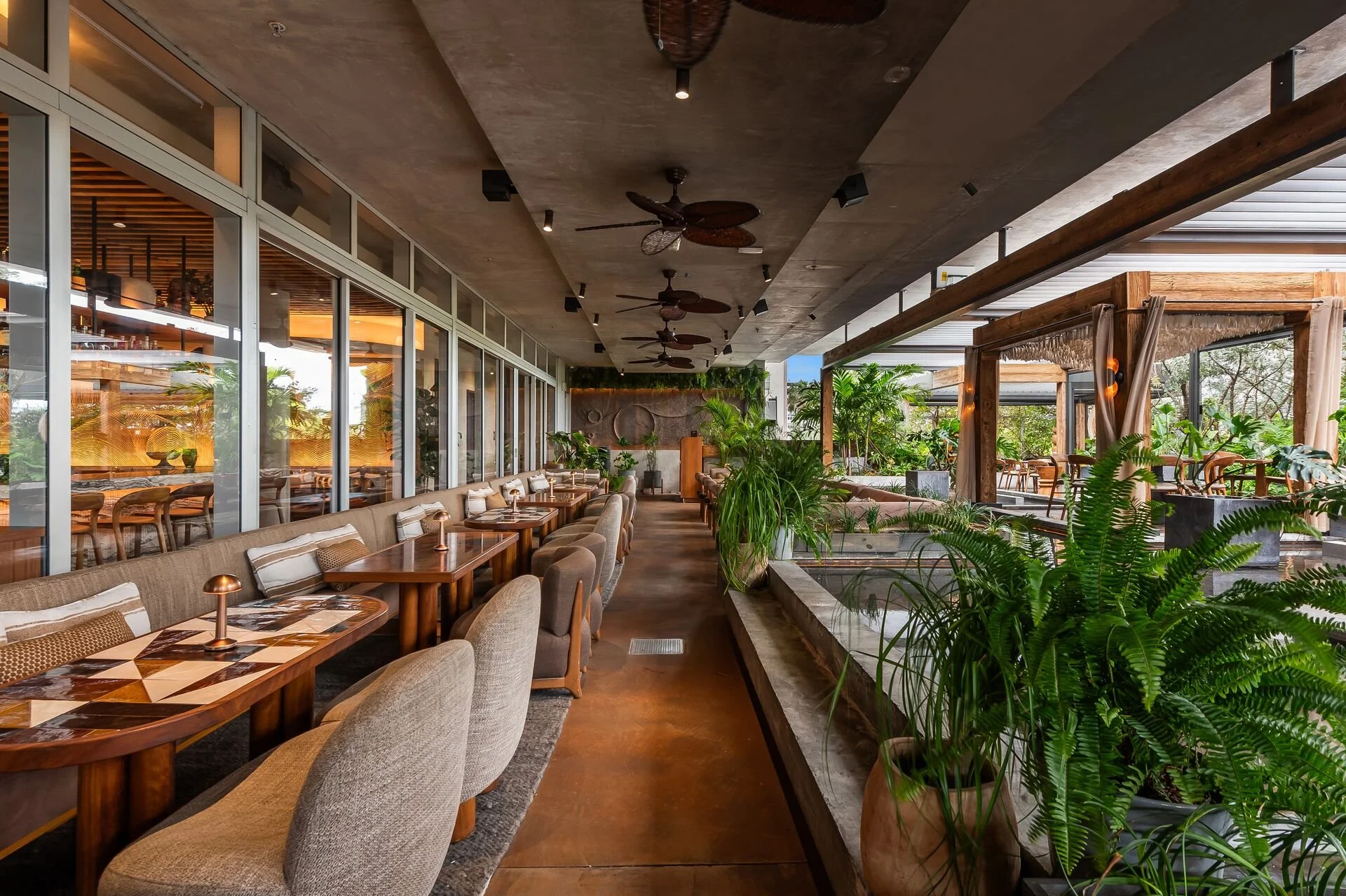 Interior of a modern restaurant with beige chairs and wooden tables, lush green plants, glass walls, and ceiling fans, blending indoor and outdoor spaces.