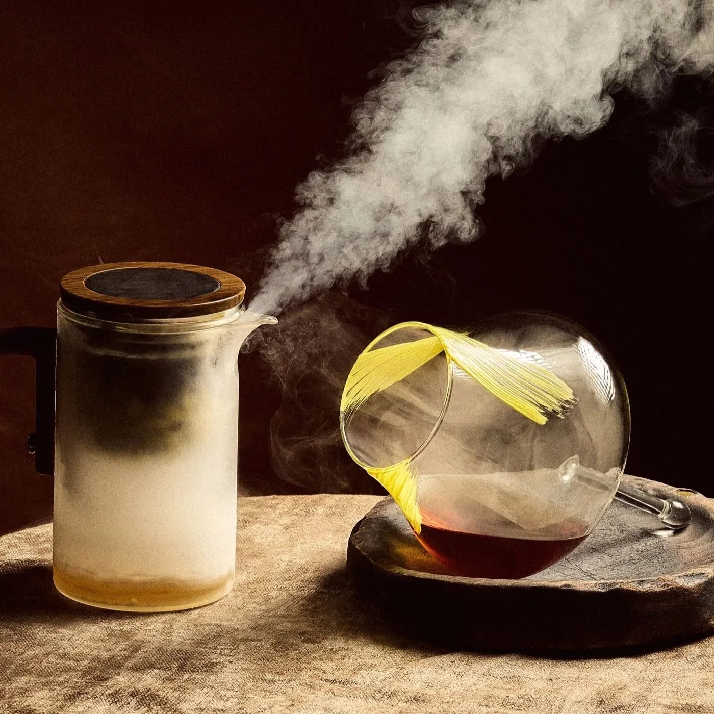 A glass of red wine, a glass teapot with steam coming out, and a clear glass with a yellow paintbrush inside, placed on a rustic wooden tray on a textured beige cloth.