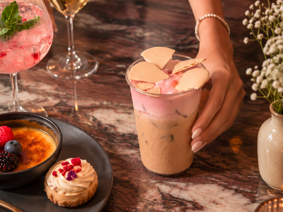 Person placing a milkshake topped with white chocolate pieces and pink whipped cream on a wooden table surrounded by drinks and desserts.