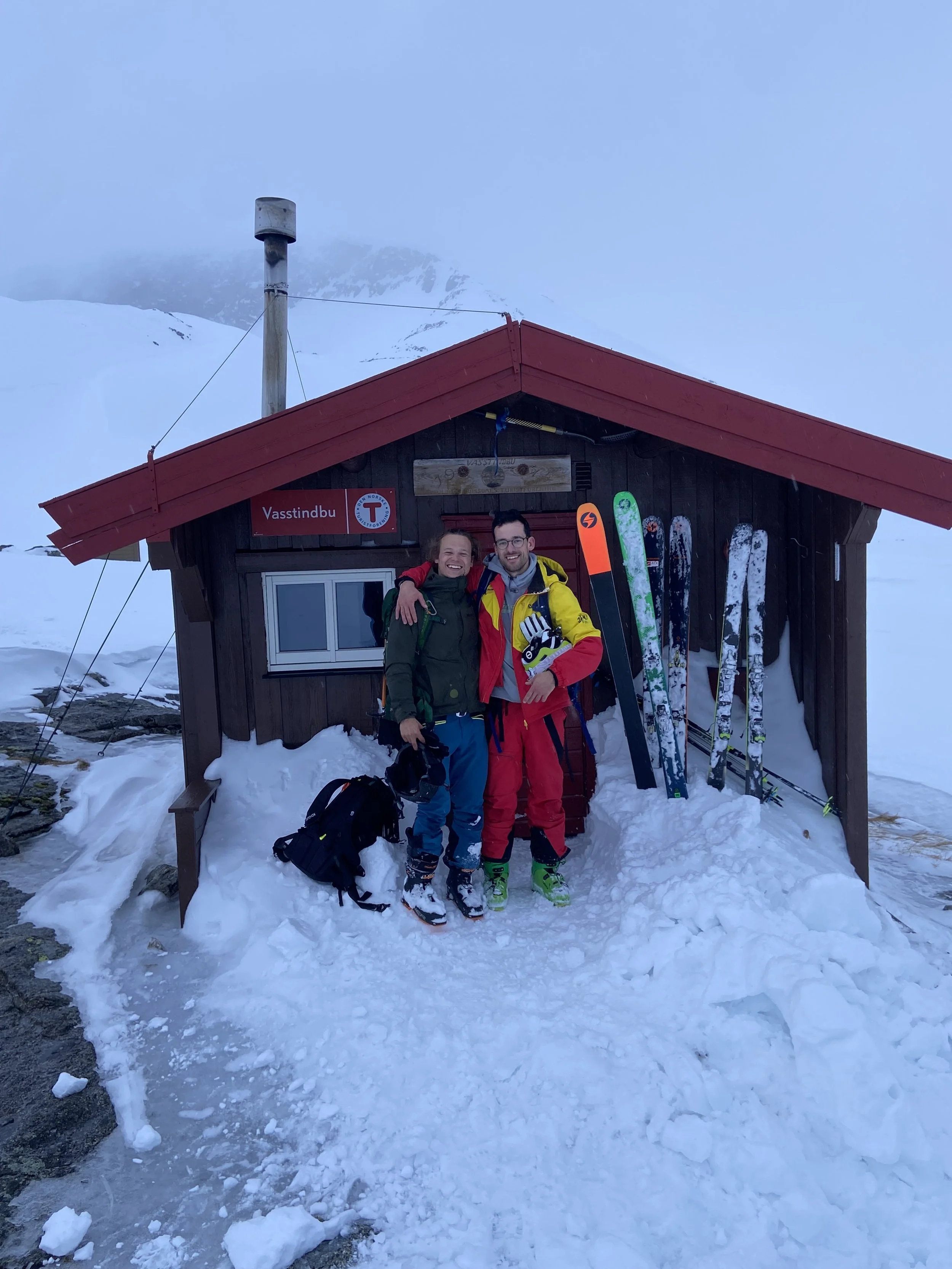 To personer står foran en liten hytte i snødekt fjellterreng, iført skiklær og med skøyter. De smiler og har armene rundt hverandre.