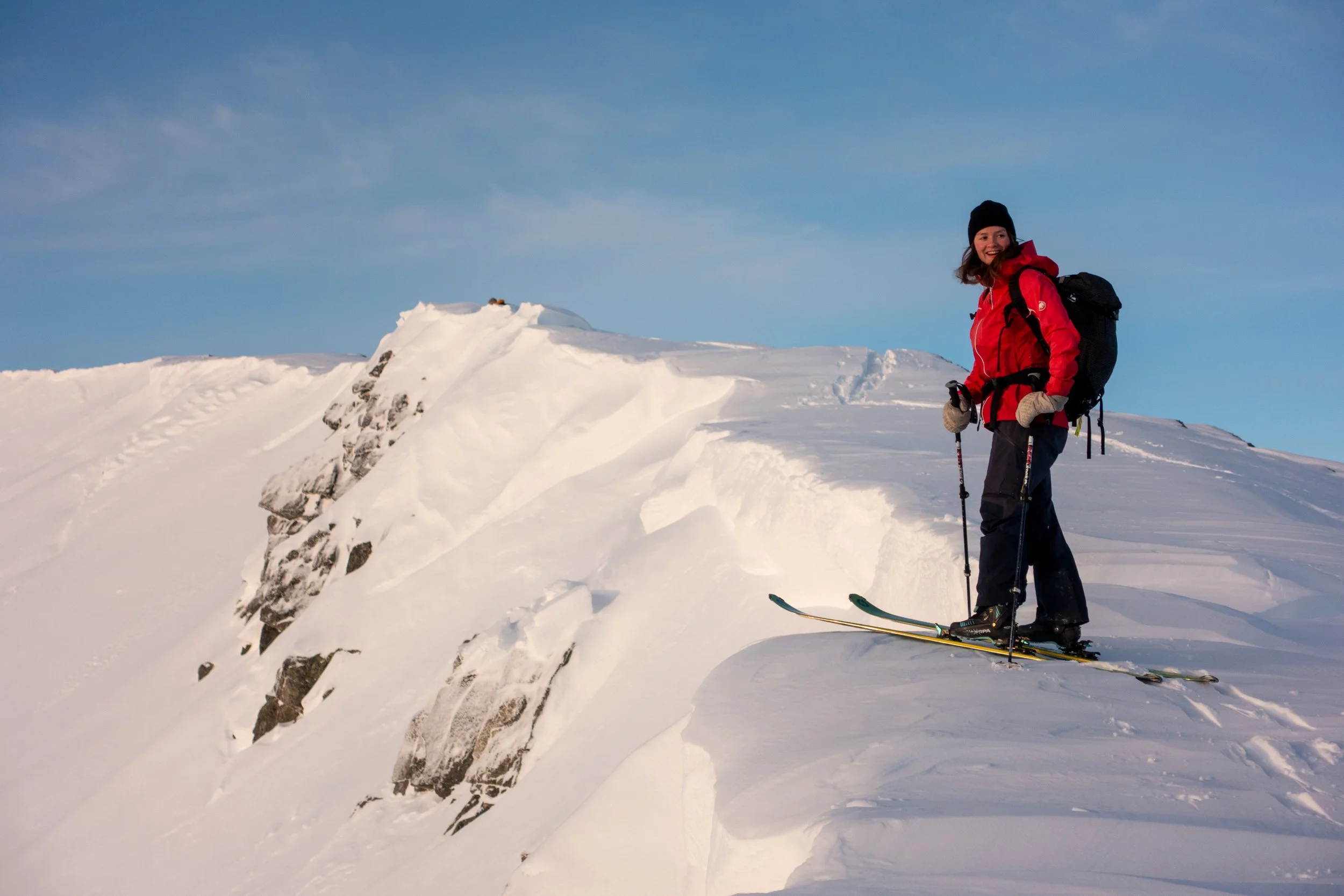 En kvinne iført rød jakke og svart hatt står på snøhop i fjellandskap med skisportutstyr og ser mot kameraet under klar himmel.