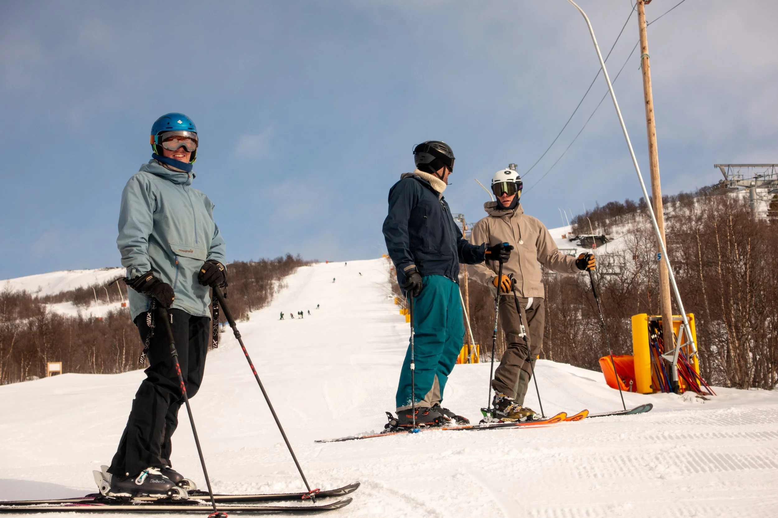 Tre personer som står på ski ved en snødekket skiheis, med bakker og andre skiløpere i bakgrunnen, under en klar himmel.