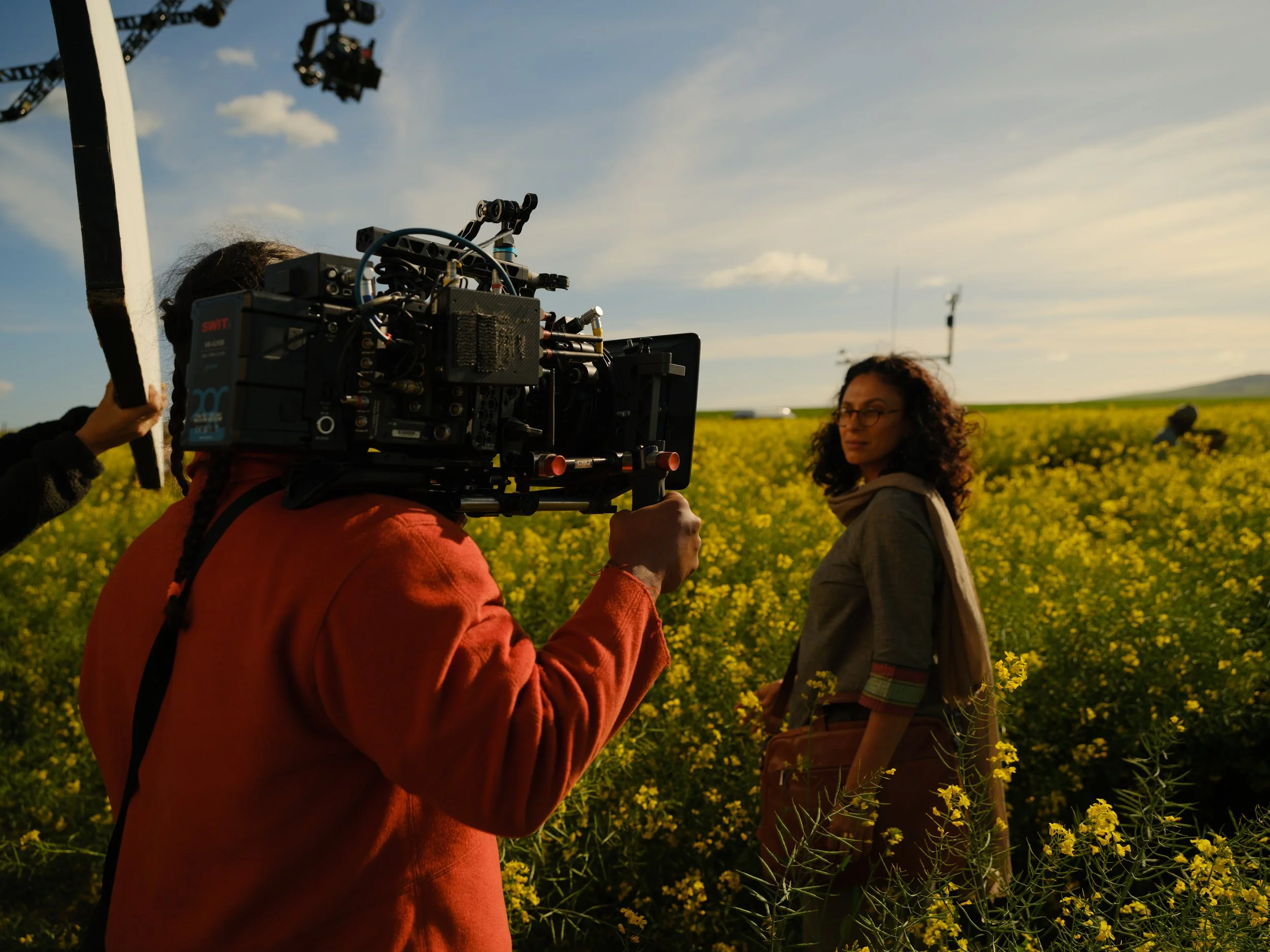 A woman stands in a field of yellow flowers during a film shoot, with a camera operator filming her. The scene is outdoors with a clear sky and some clouds.