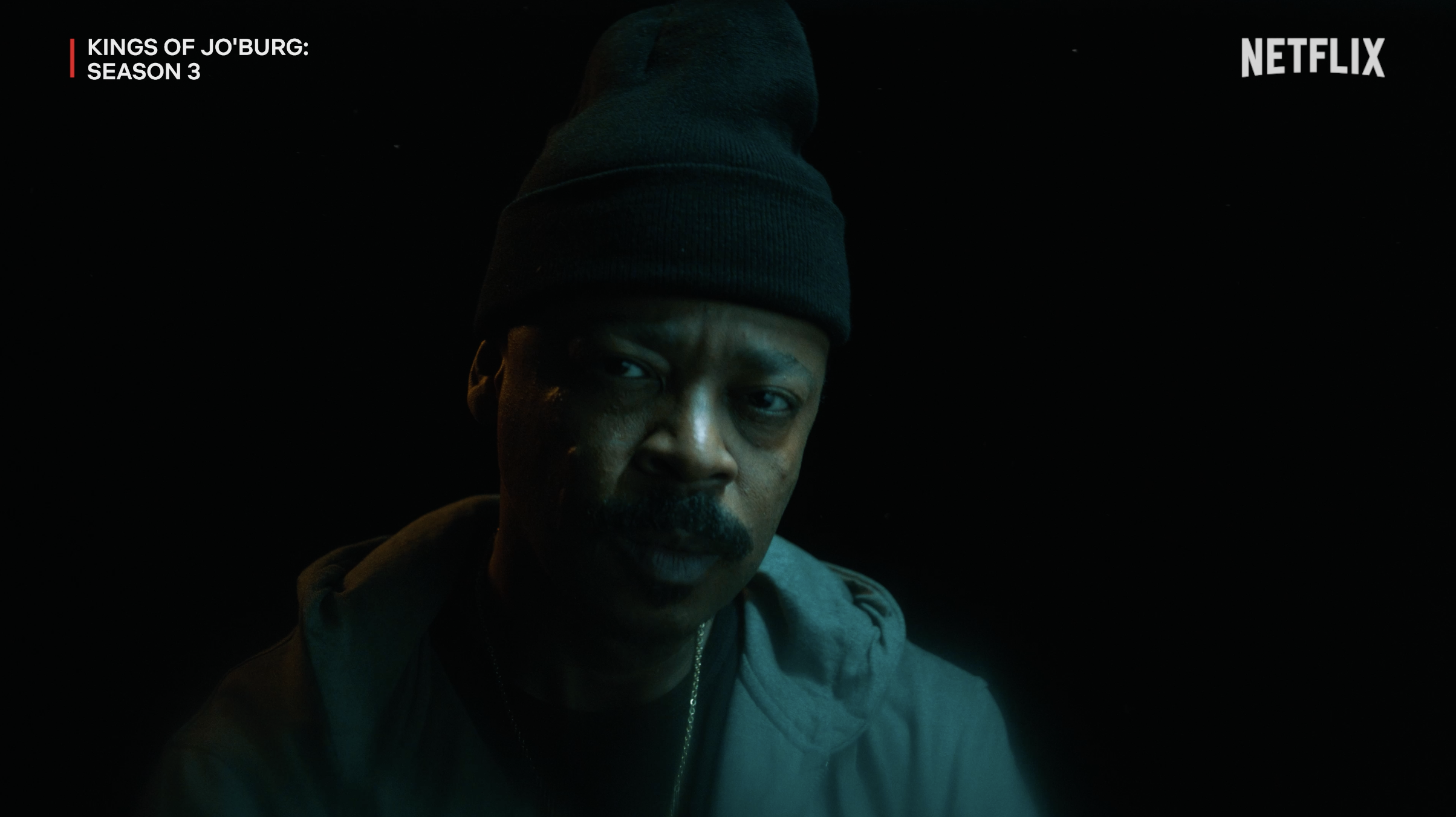 A man wearing a black beanie and gray hoodie, looking serious, with a dark background. Text overlay includes 'Kings of Jo'burg: Season 3' and the Netflix logo.