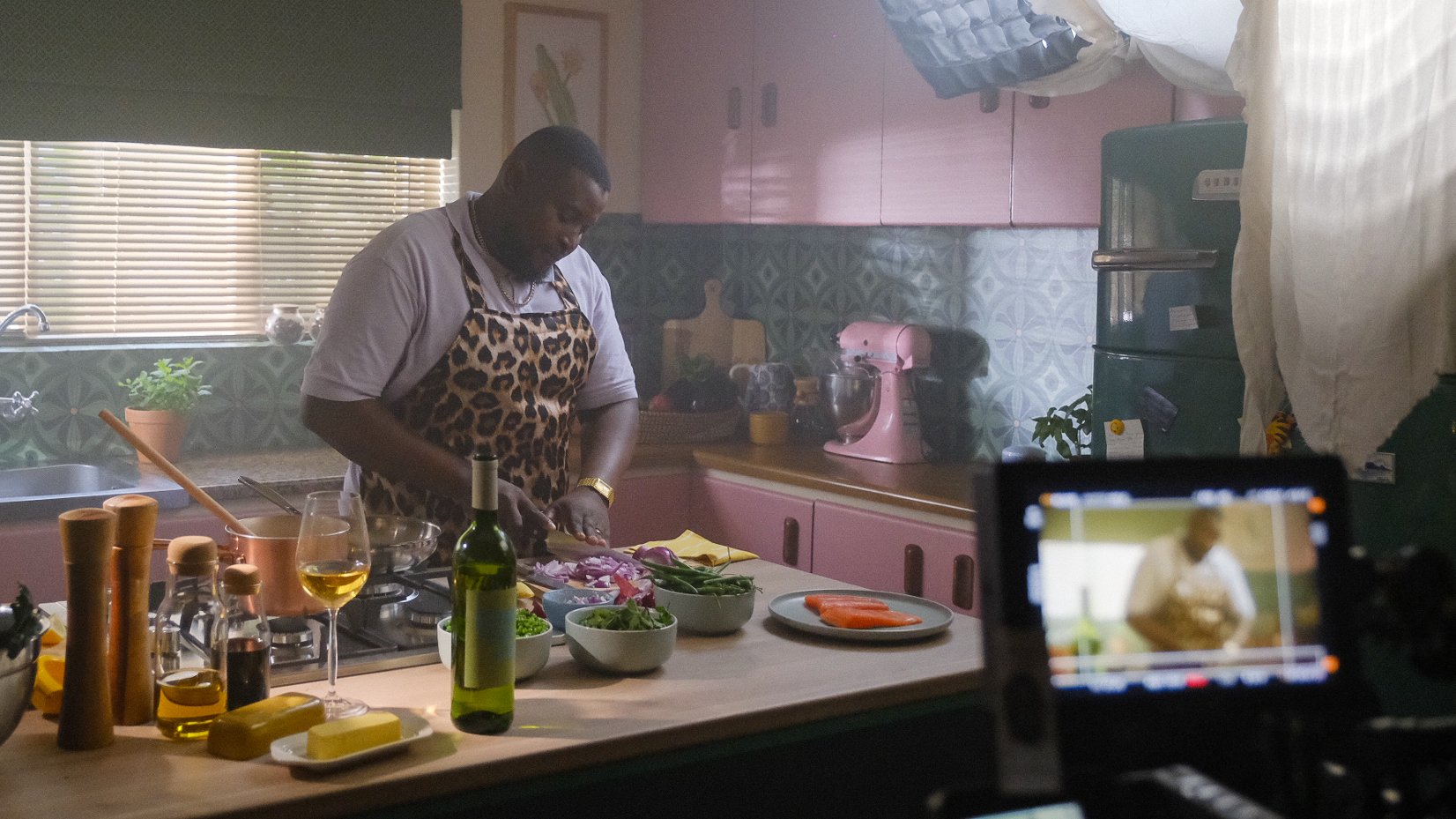 A man wearing a leopard print apron preparing food in a kitchen with pink cabinets, various bowls of vegetables, a bottle of wine, and a glass of white wine on the counter. There is a camera filming him.