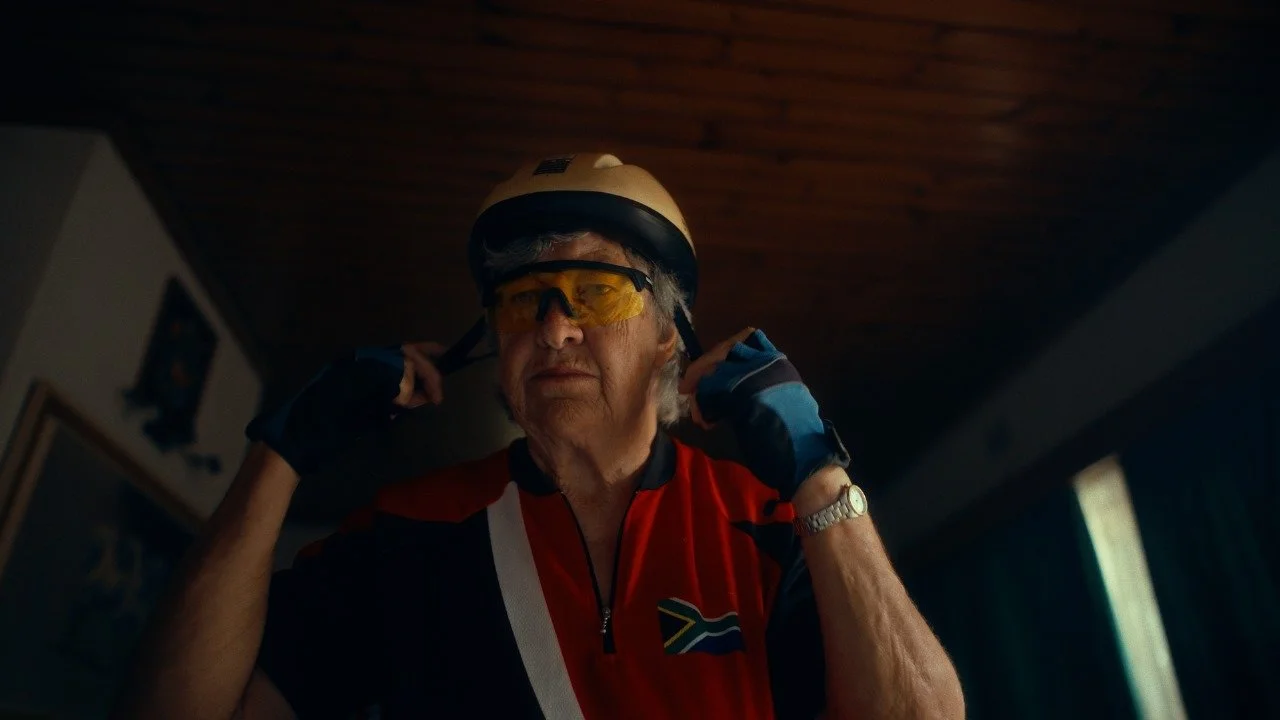 An elderly man wearing a cycling helmet, tinted sports glasses, a red cycling jersey with South African flag, and fingerless gloves, adjusting his helmet indoors near a window.