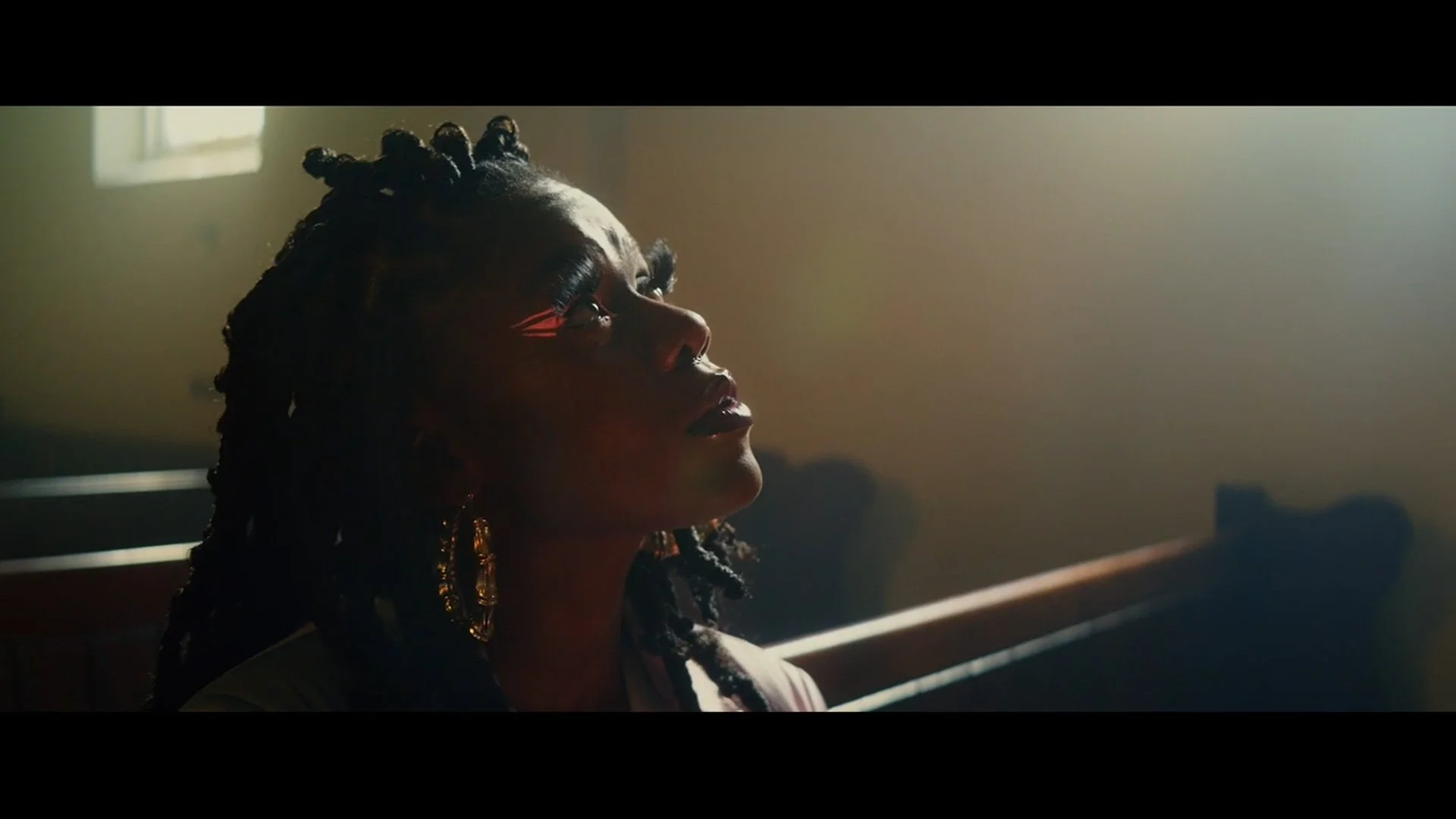 Woman with braided hair and earrings sitting on a bench in a room with sunlight coming through the window.